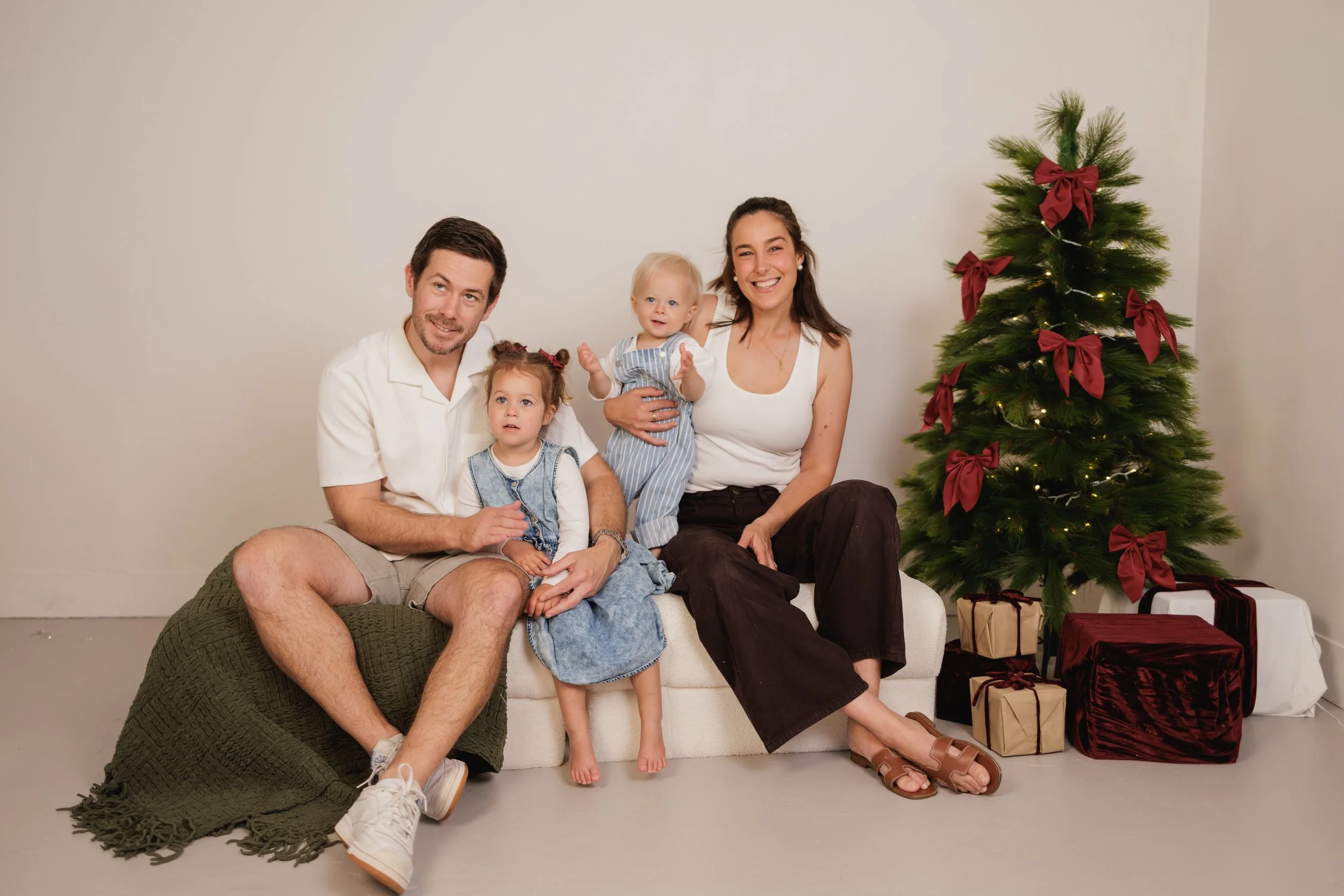 A family of four sitting on a white couch near a decorated Christmas tree with gift boxes underneath, against a plain white wall.