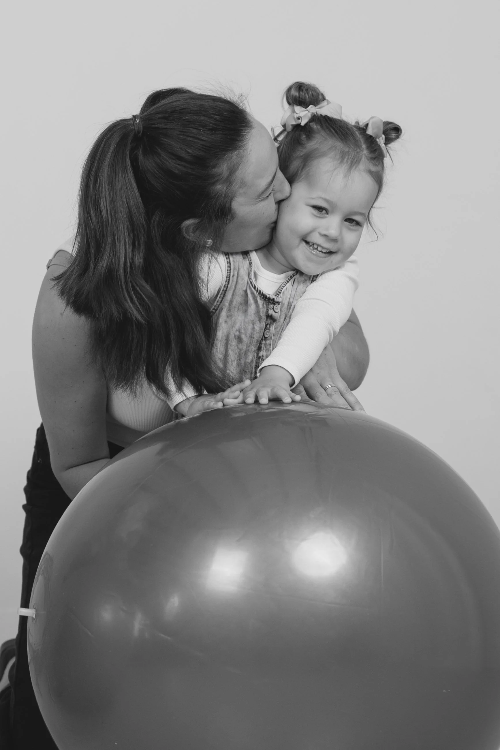 A woman with long hair in a ponytail kissing a smiling young girl with two topknot hair buns and ribbon bows on her head, sitting behind large exercise ball.