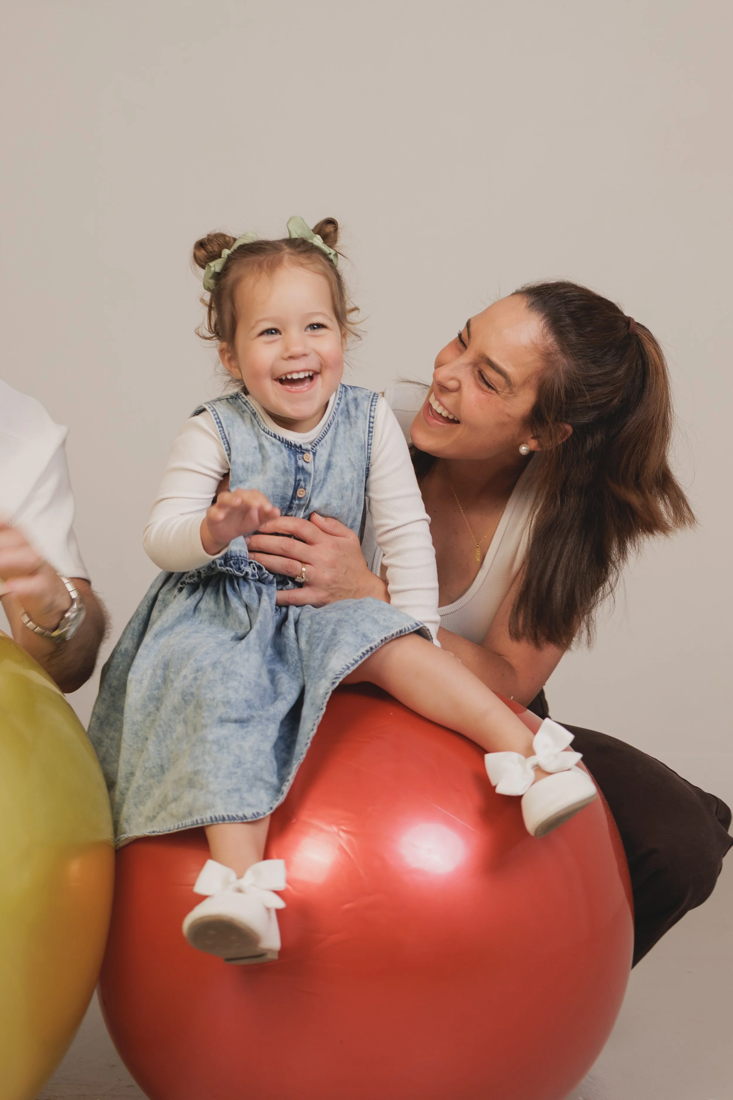 A woman and a young girl sitting on a large red balloon, smiling and laughing, with a plain background.