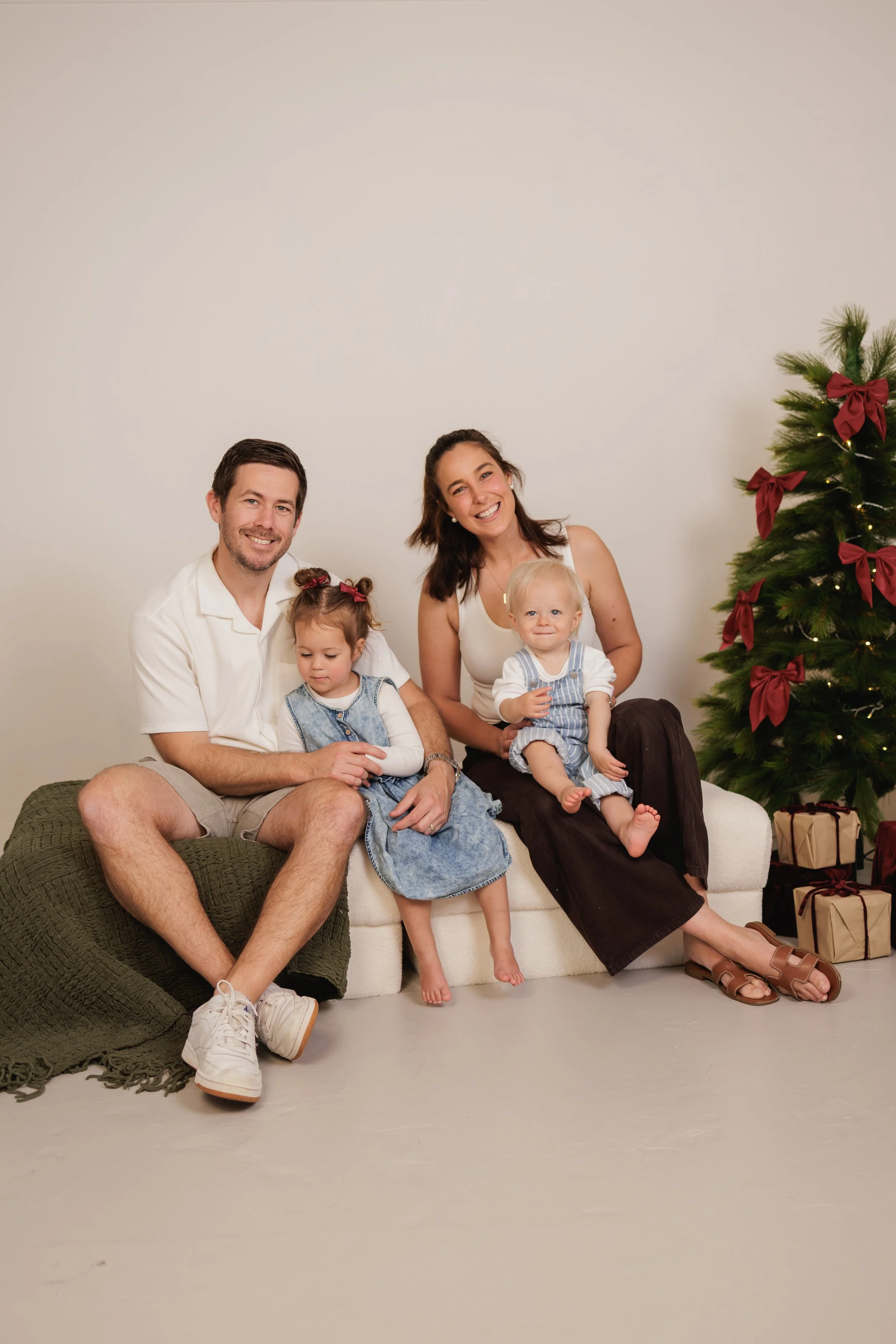 A family of four sitting on a white couch next to a decorated Christmas tree with red bows and lights, all smiling and celebrating together.