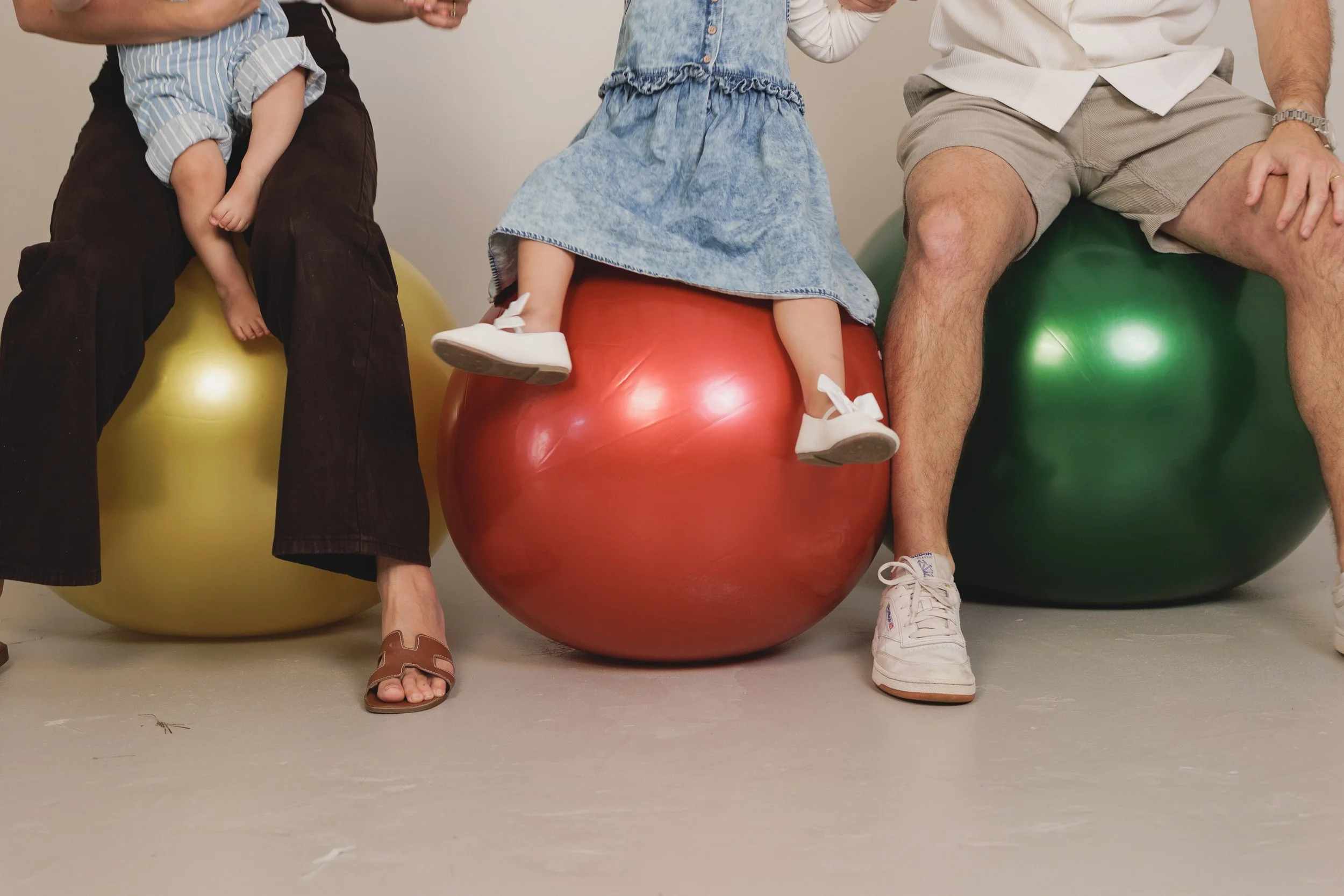 Three people sitting on large exercise balls, showing their legs and feet. The person on the left is barefoot with toe rings, the middle person is wearing white sneakers, and the right person is wearing white sneakers with purple accents. They are in