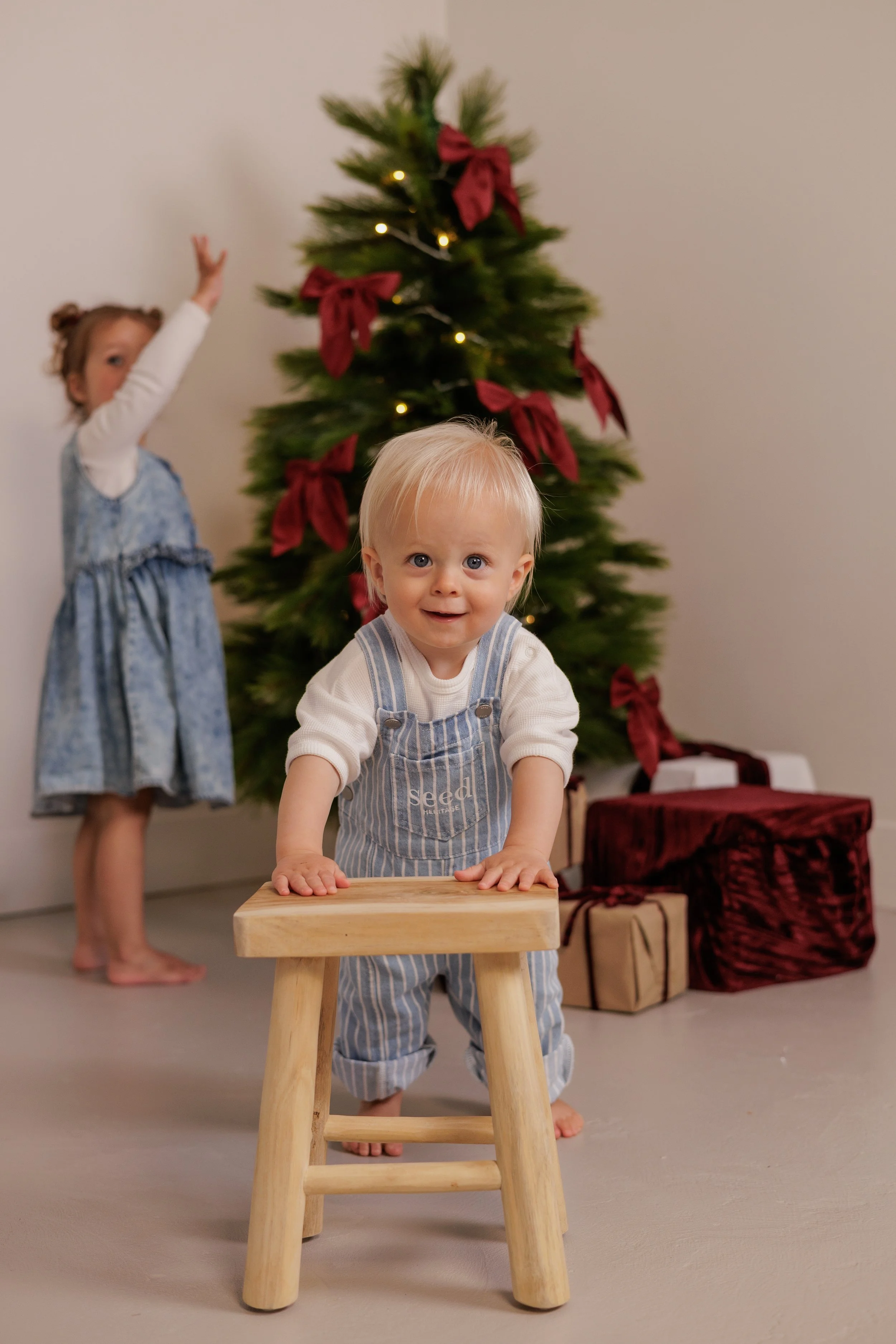 Two young children, a boy and a girl, decorating a Christmas tree with red ribbons and fairy lights. The boy, with blond hair, is standing in front of a small wooden stool, smiling at the camera. The girl, with curly hair in pigtails, is reaching up 
