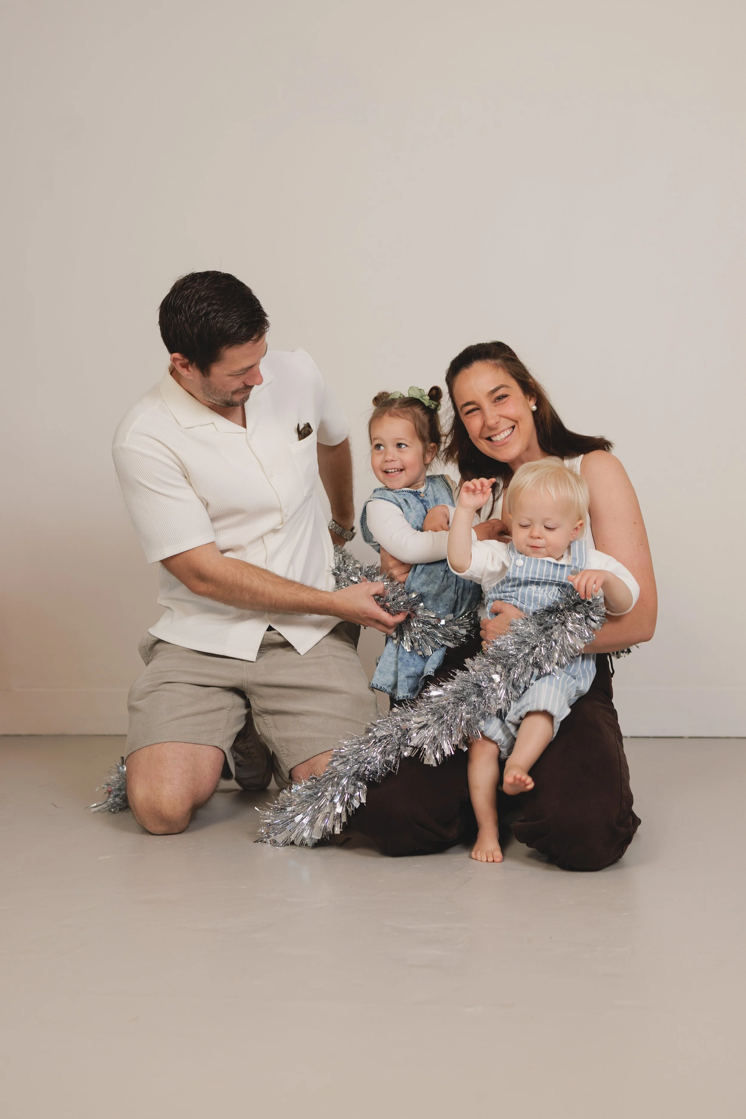 A woman holding two children, one boy and one girl, with a man kneeling next to them, all smiling and wrapping tinsel around them against a plain beige wall.