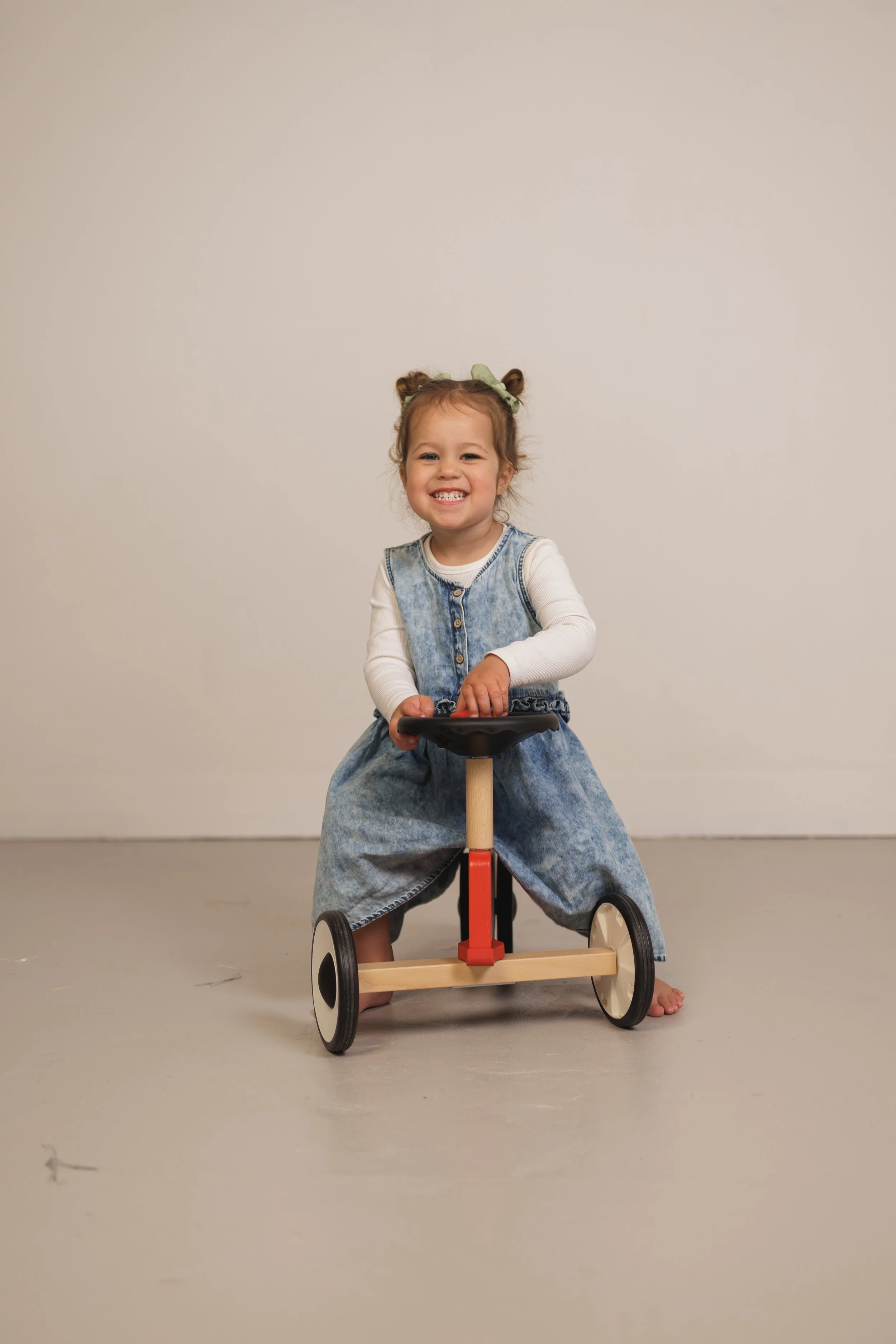 A young girl with curly hair tied in two buns, wearing a denim dress and a long sleeve white shirt, smiling and sitting on a small wooden and red tricycle, against a plain light-colored wall.