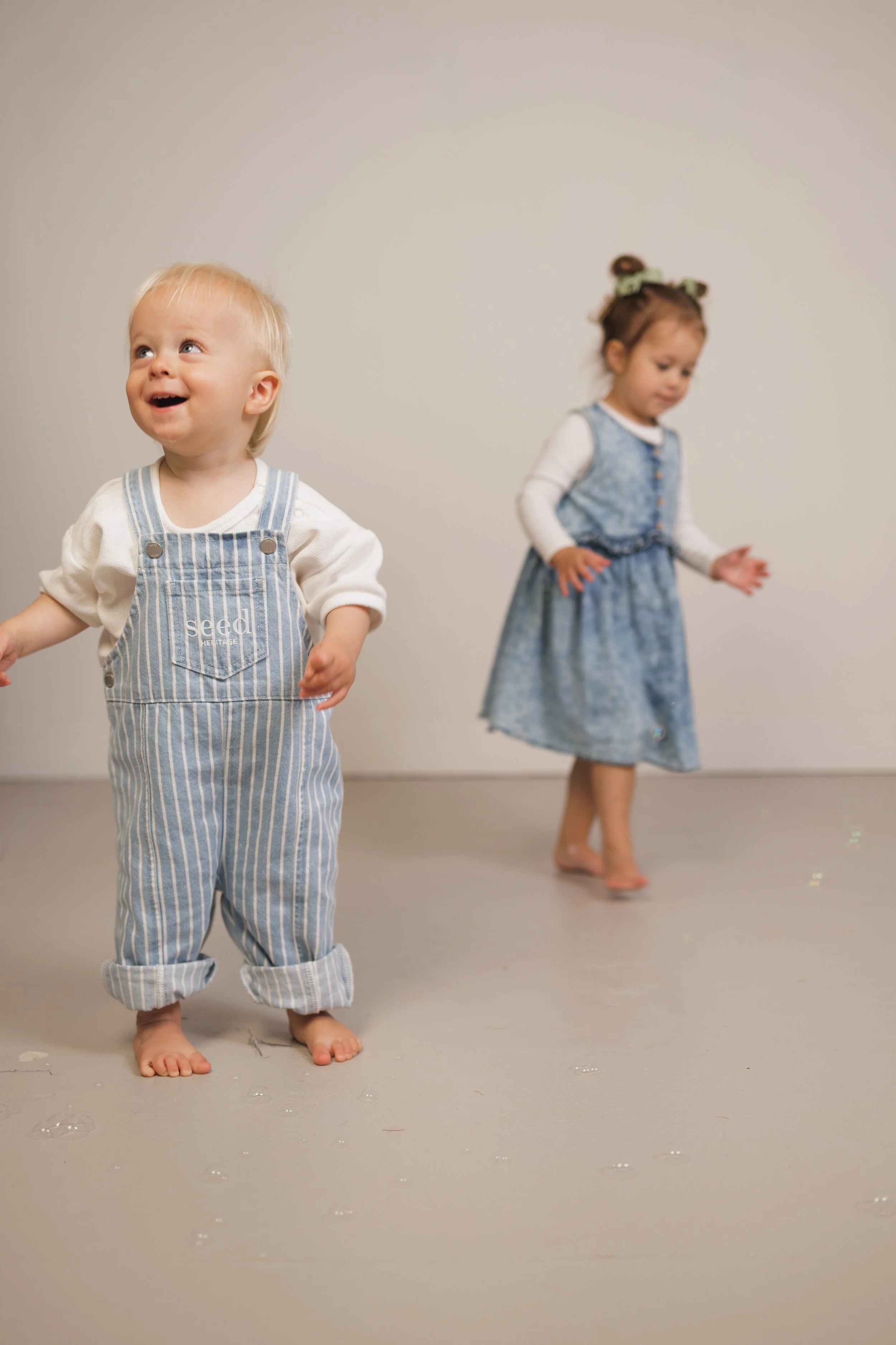 Two young children, one boy and one girl, are standing barefoot on a gallery floor, with some soap bubbles scattered around them. The boy, with blonde hair and wearing striped overalls, is smiling and looking upwards. The girl, with brown hair styled