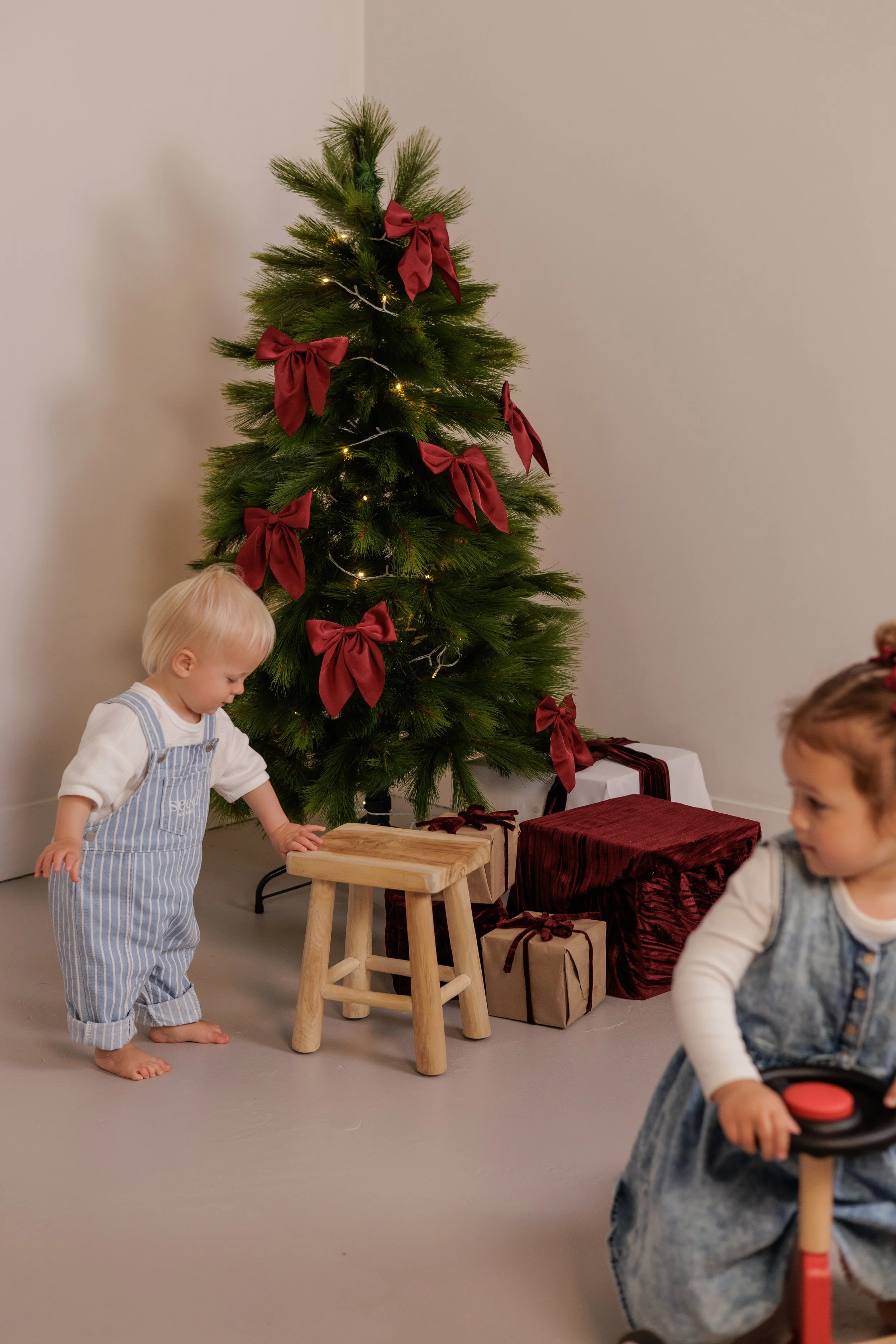 Children playing near a Christmas tree decorated with red bows, with wrapped gifts underneath, in a cozy indoor setting.