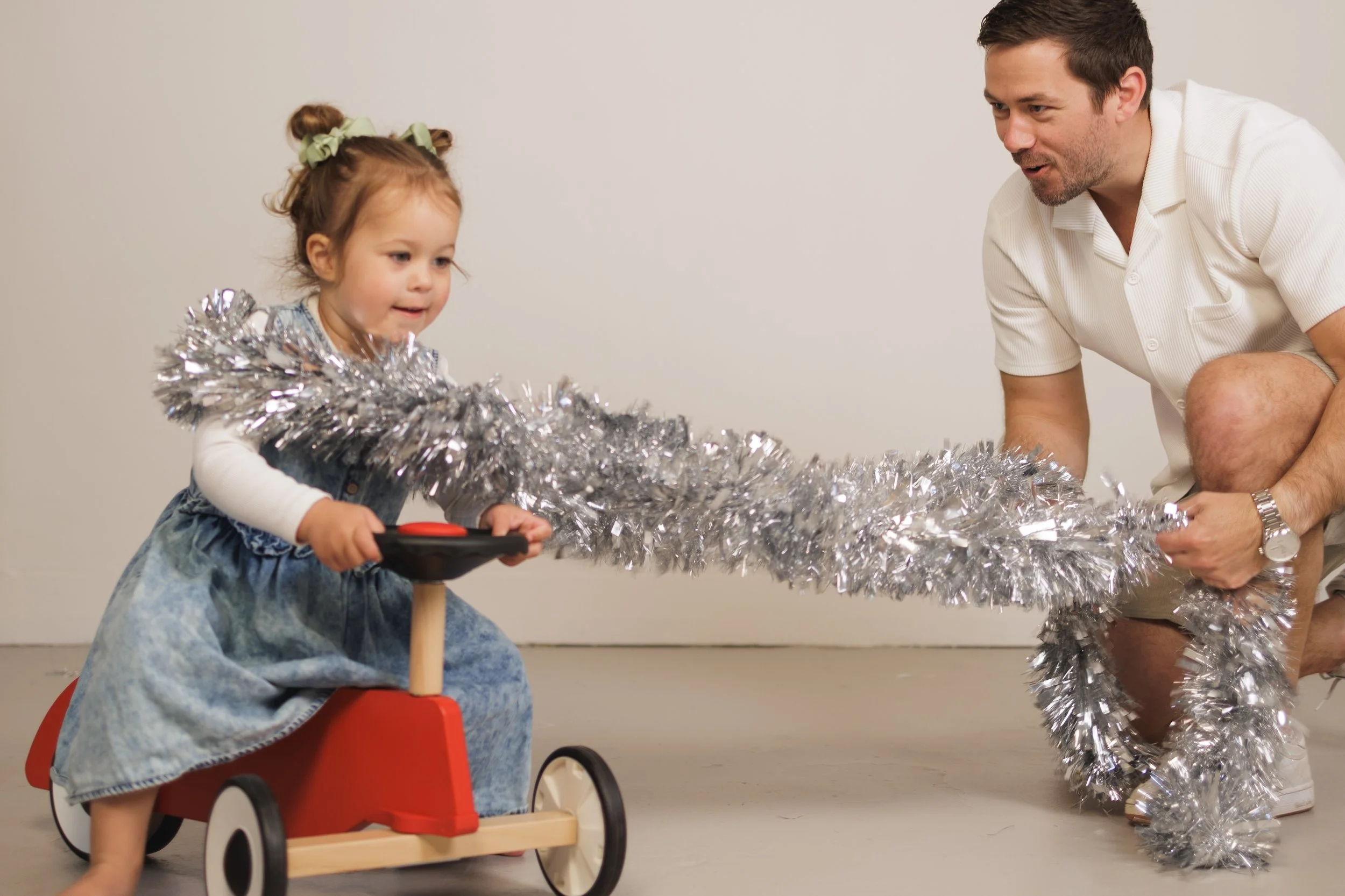 A young girl riding a red and wooden tray ride with black wheels, holding a black steering wheel, is being helped by a man to wrap silver tinsel around her. The girl has light brown hair with two green bows, and is wearing a denim dress with a white 
