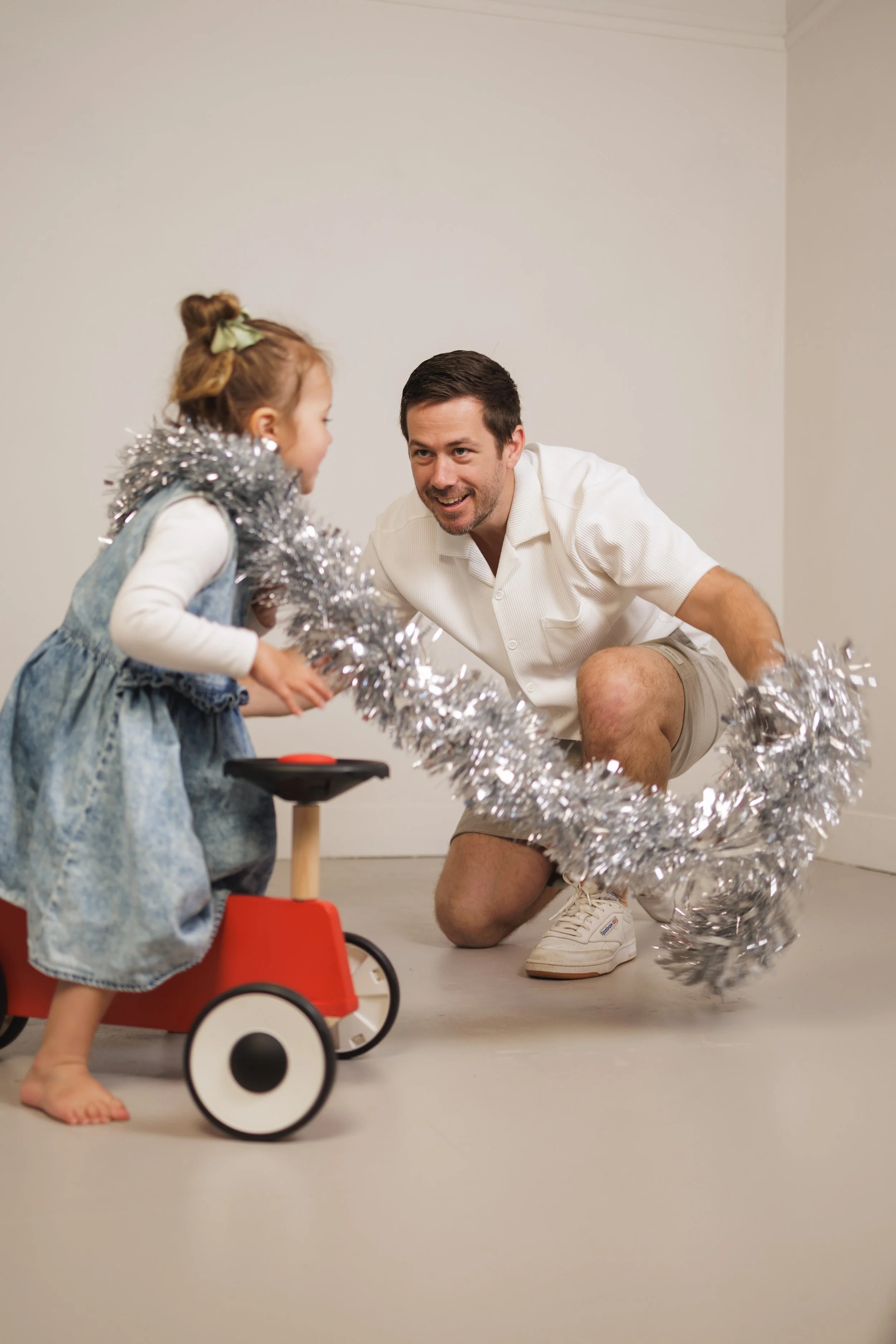 A man crouching down and smiling at a young girl who is engaged in a playful activity with silver tinsel. The girl is wearing a blue dress with a bow in her hair and is standing next to a red gasoline-powered toy tricycle. The scene appears to be ind