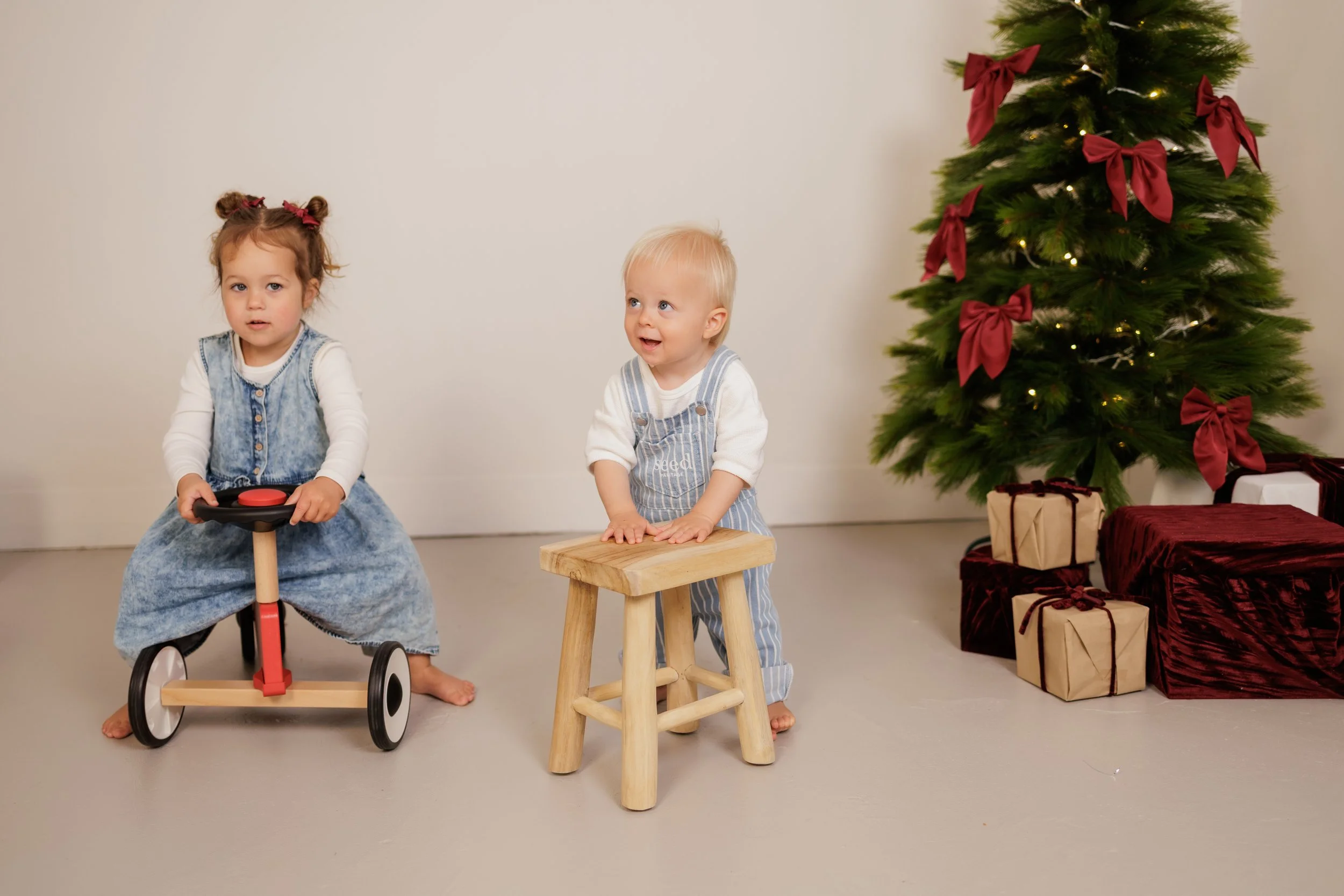 Two young children, a girl on a scooter and a boy on a small wooden stool, are in a room decorated for Christmas with a Christmas tree and wrapped presents in the background.