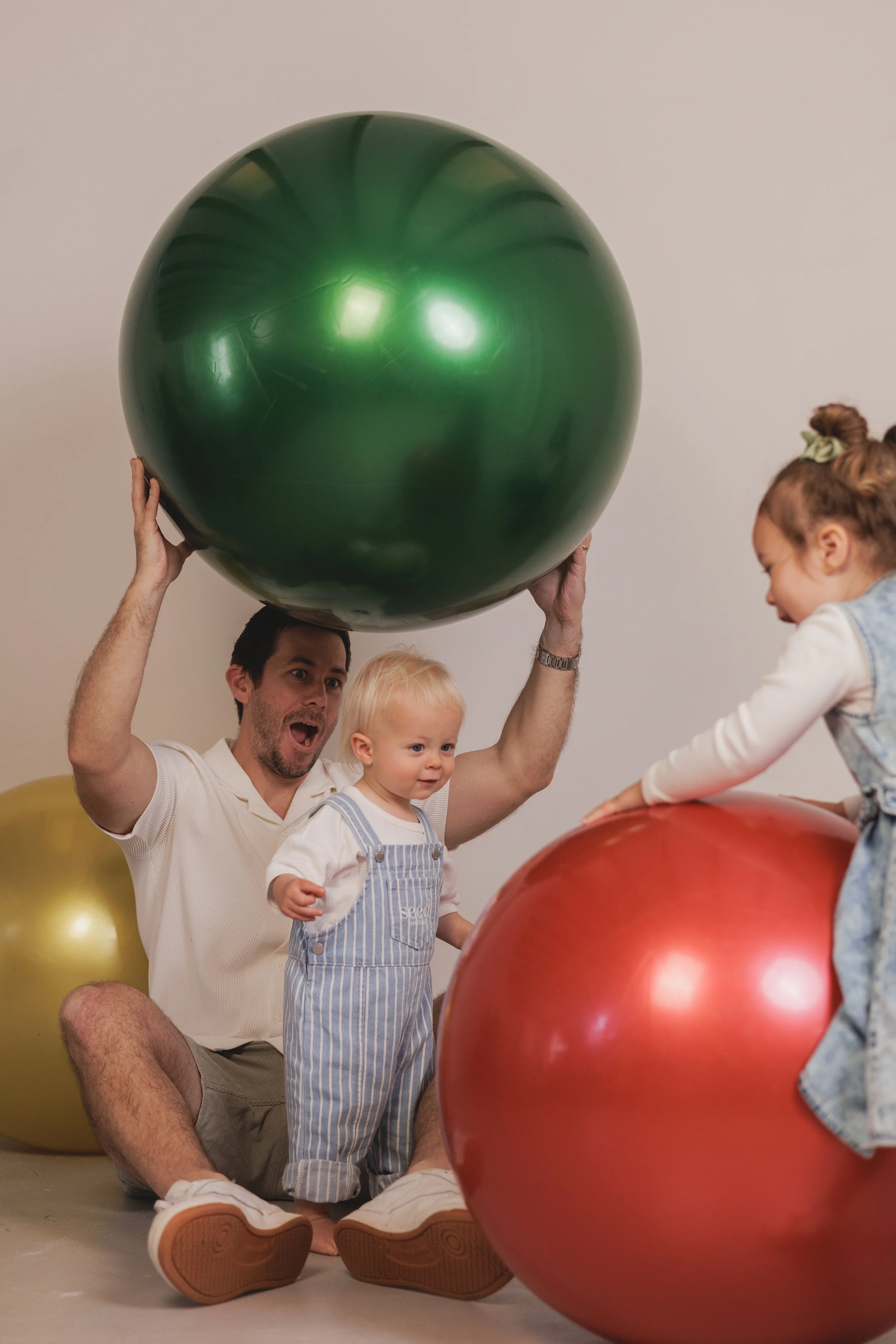 A man, two young children, and a girl playing with large red, green, and gold exercise balls indoors against a plain wall.