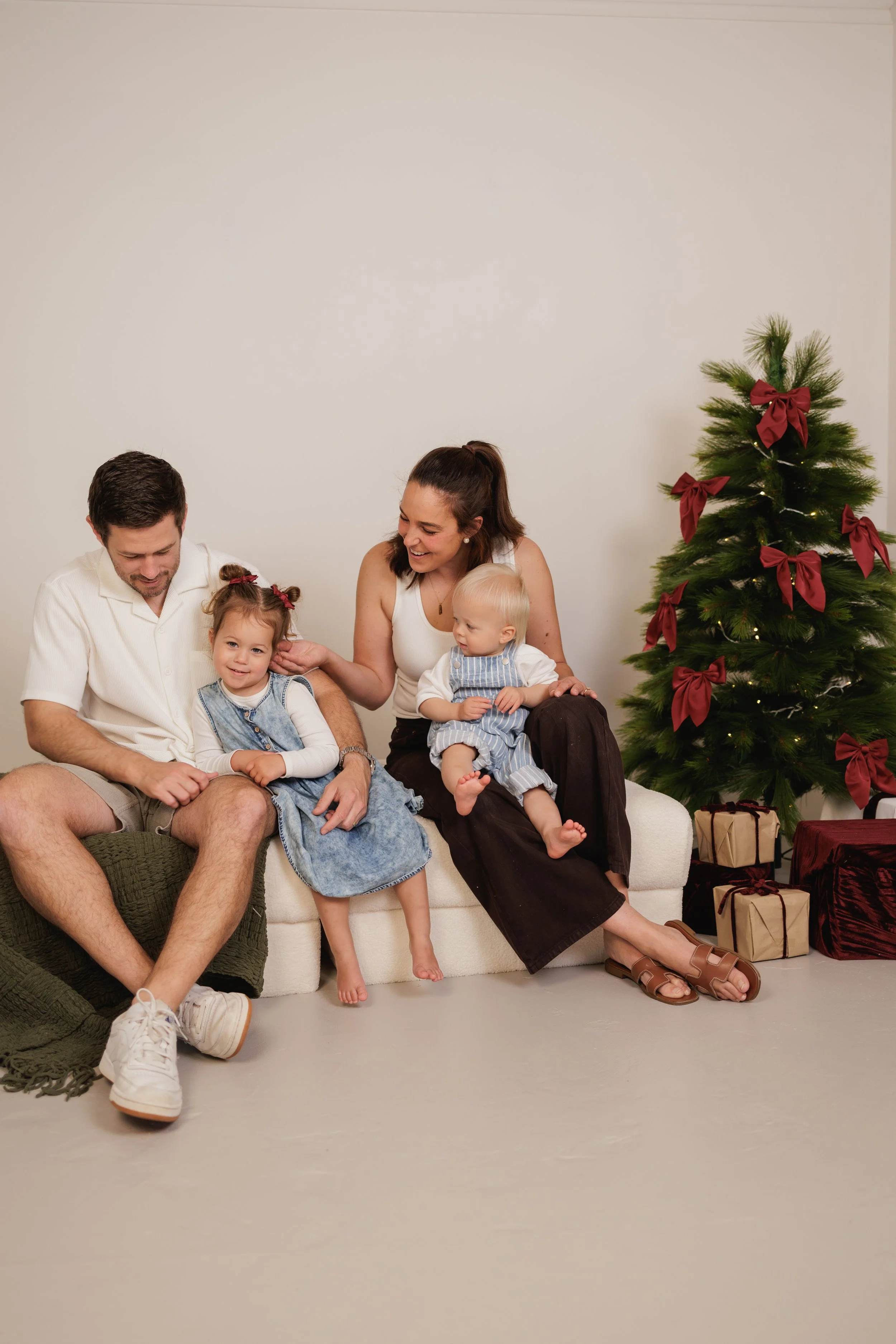 A family of four, including two young children, sitting on a white sofa near a Christmas tree decorated with red bows and surrounded by wrapped gifts. The family is smiling and appears to be enjoying a holiday moment.