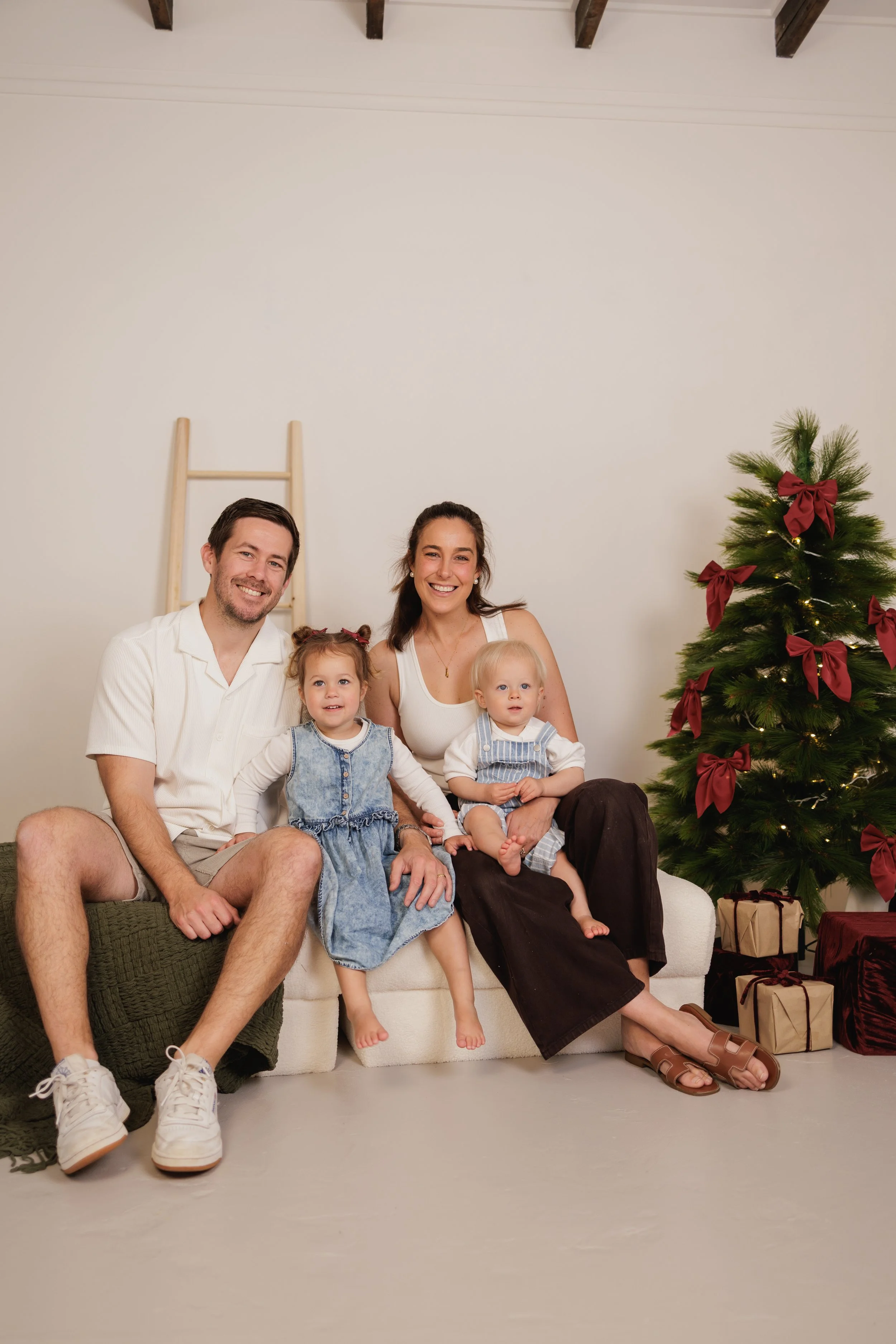 A family of four with two young children sitting on a beige sofa in a festive setting. A Christmas tree decorated with red bows and gift packages on the floor are visible. The family appears happy and relaxed, celebrating the holiday season.