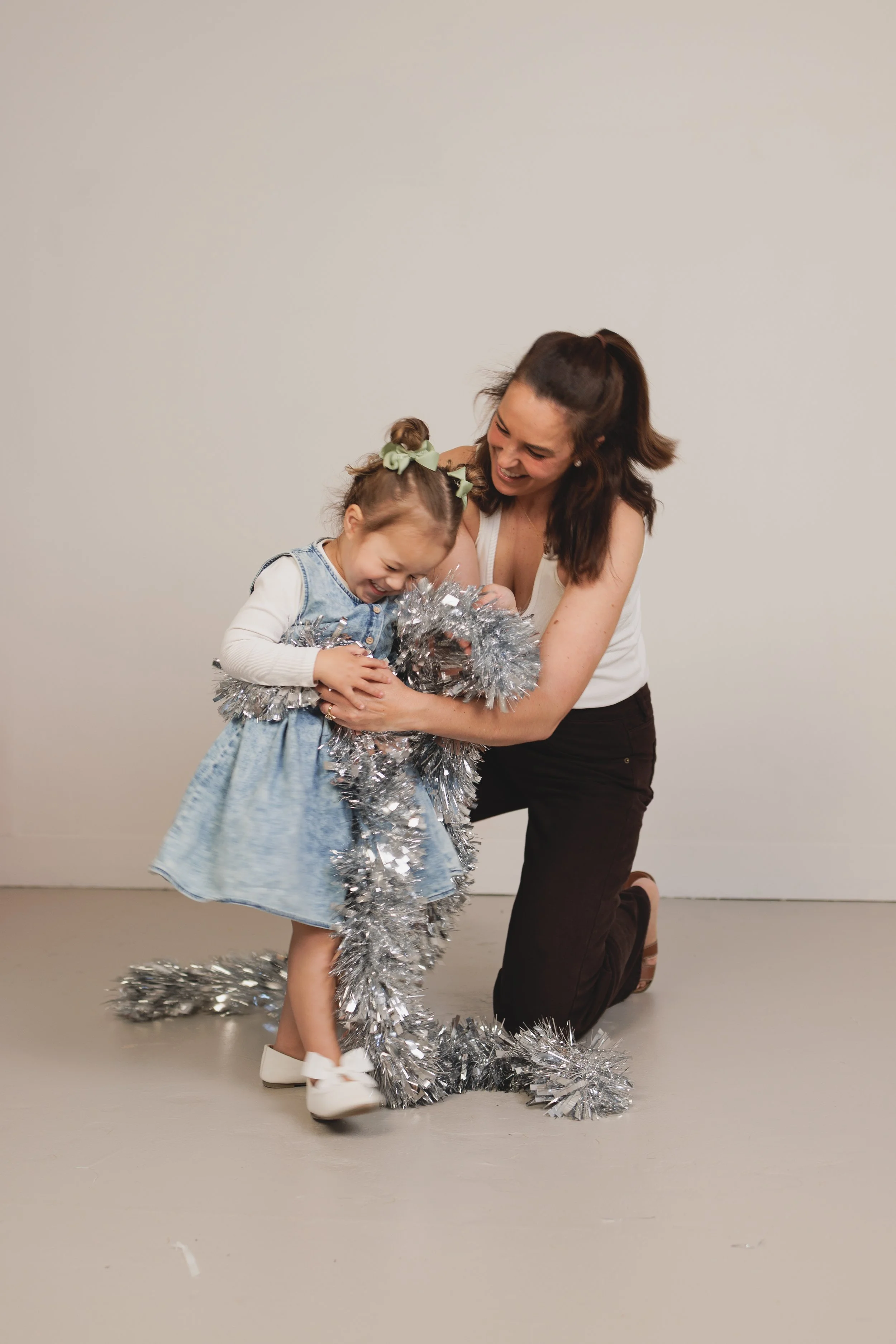 A woman and a young girl happily playing with silver tinsel in a plain room with a light-colored floor and wall.