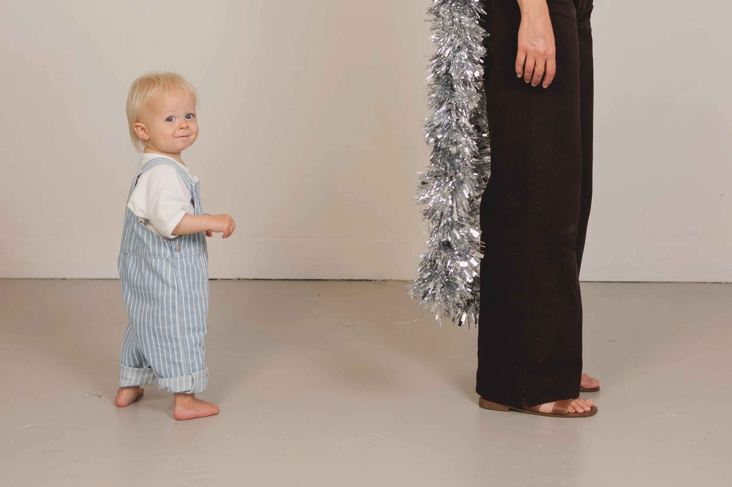 A small child with blonde hair and blue eyes, wearing light blue and white striped overalls, standing barefoot and looking up at an adult. The adult, partially visible, is wearing dark pants and sandals with a gold ring on their finger. They are hold
