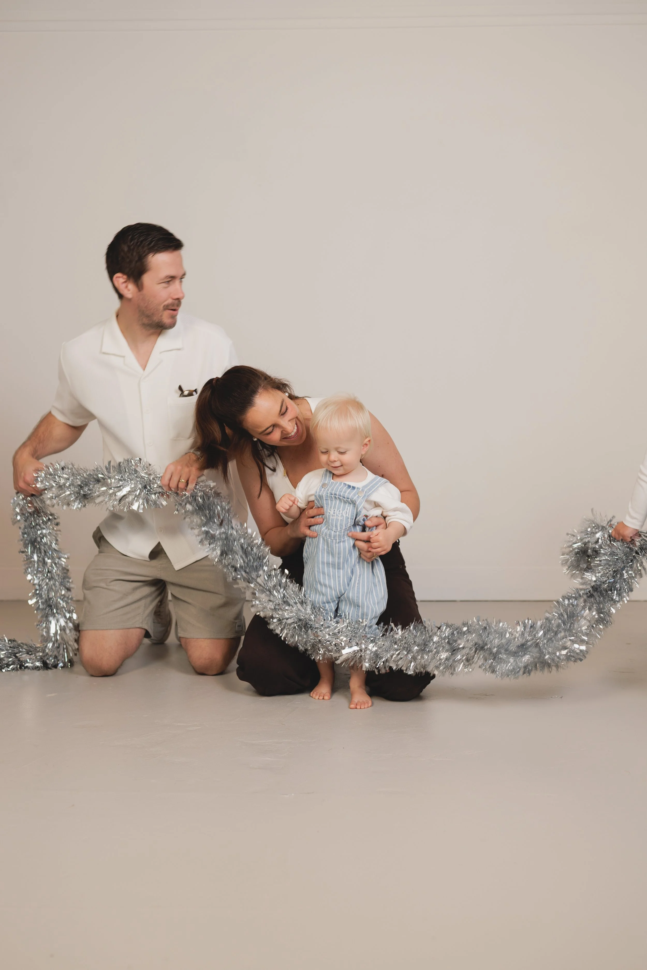 A family gathered around a silver tinsel ring, celebrating during the holiday season. The woman is holding a young child, and a man is kneeling next to them. They are all smiling and enjoying a festive moment.