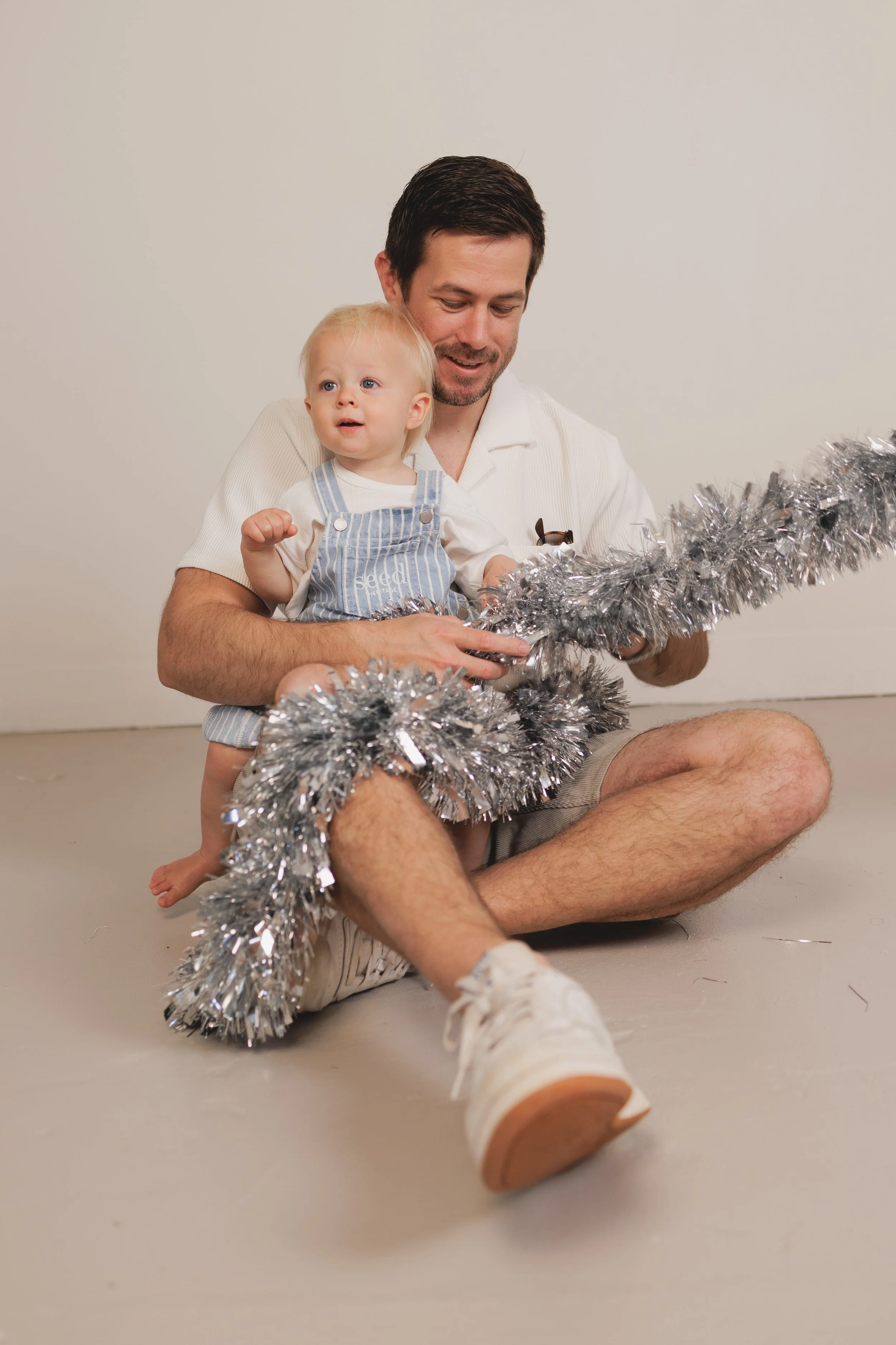 A man and a young girl sitting on the floor, holding silver tinsel, with the girl looking surprised or curious, and both appearing to enjoy a festive moment.