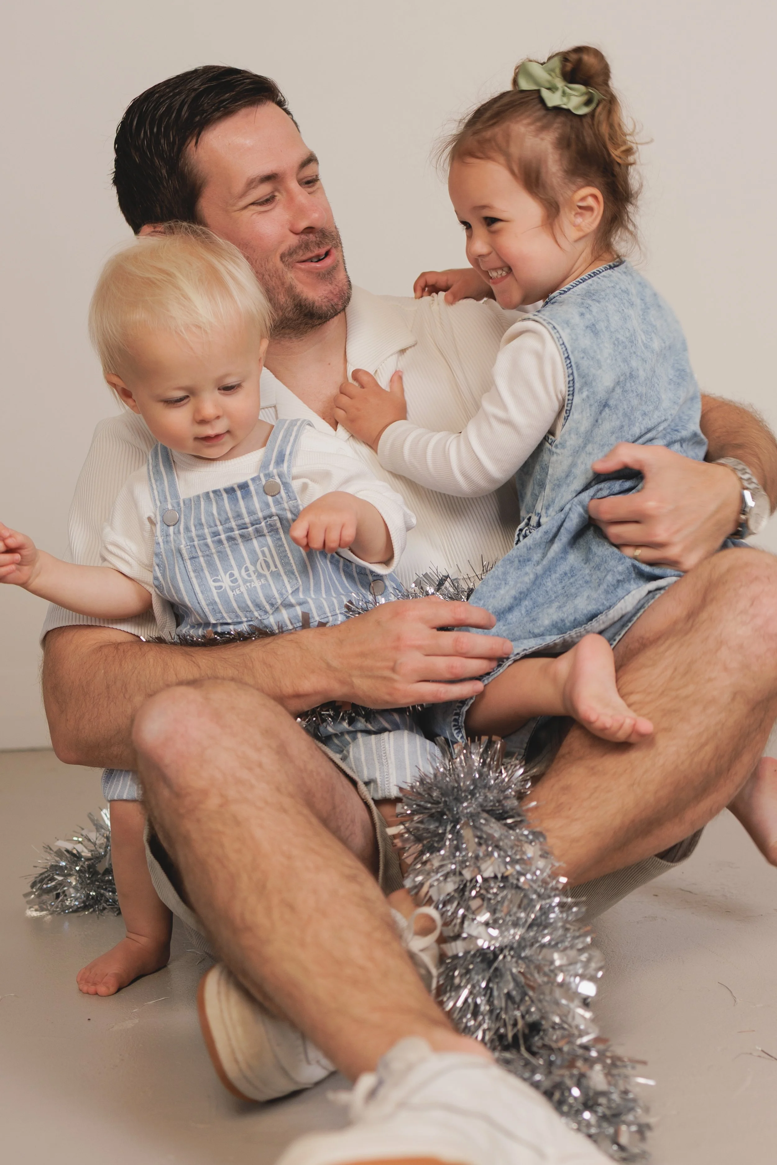 Father holding two children, a boy and a girl, as they laugh and play with silver tinsel, against a plain background.