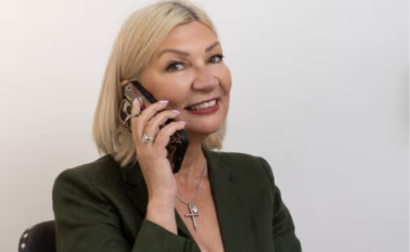 Smiling blonde woman in green blazer talking on a cell phone, wearing a cross necklace and ring, against a white background.