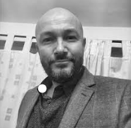 A man with a beard and a shaved head, wearing a suit jacket, standing in front of a patterned backdrop.