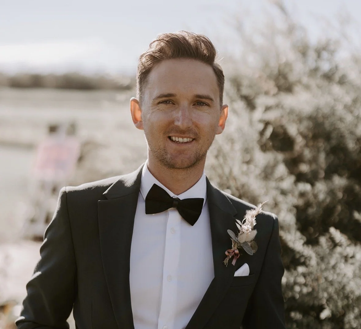 A young man dressed in a tuxedo and bow tie, smiling outdoors with trees in the background.