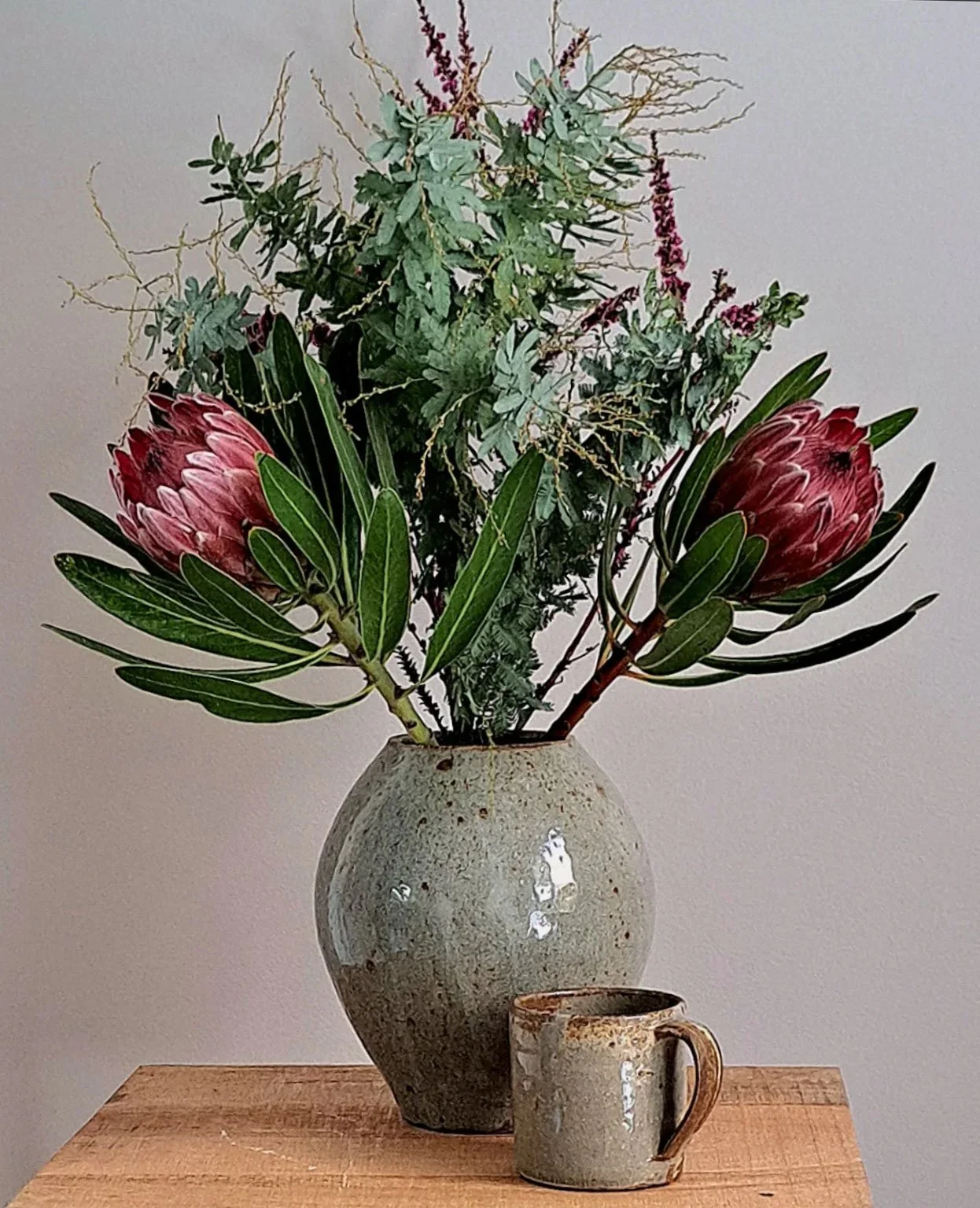 A bouquet of lush greenery and large pink protea flowers in a ceramic vase on a wooden table, with a rustic mug nearby.