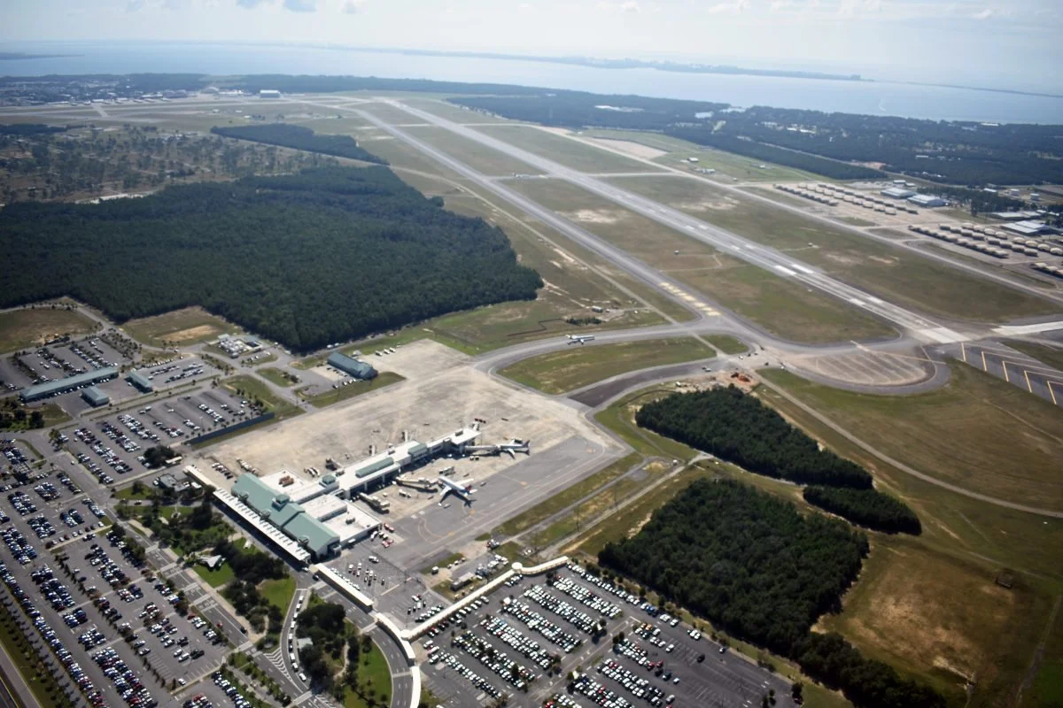 Aerial view of an airport showing runways, taxiways, a terminal building with airplanes parked, large parking lots, and surrounding green areas with trees and water in the distance.