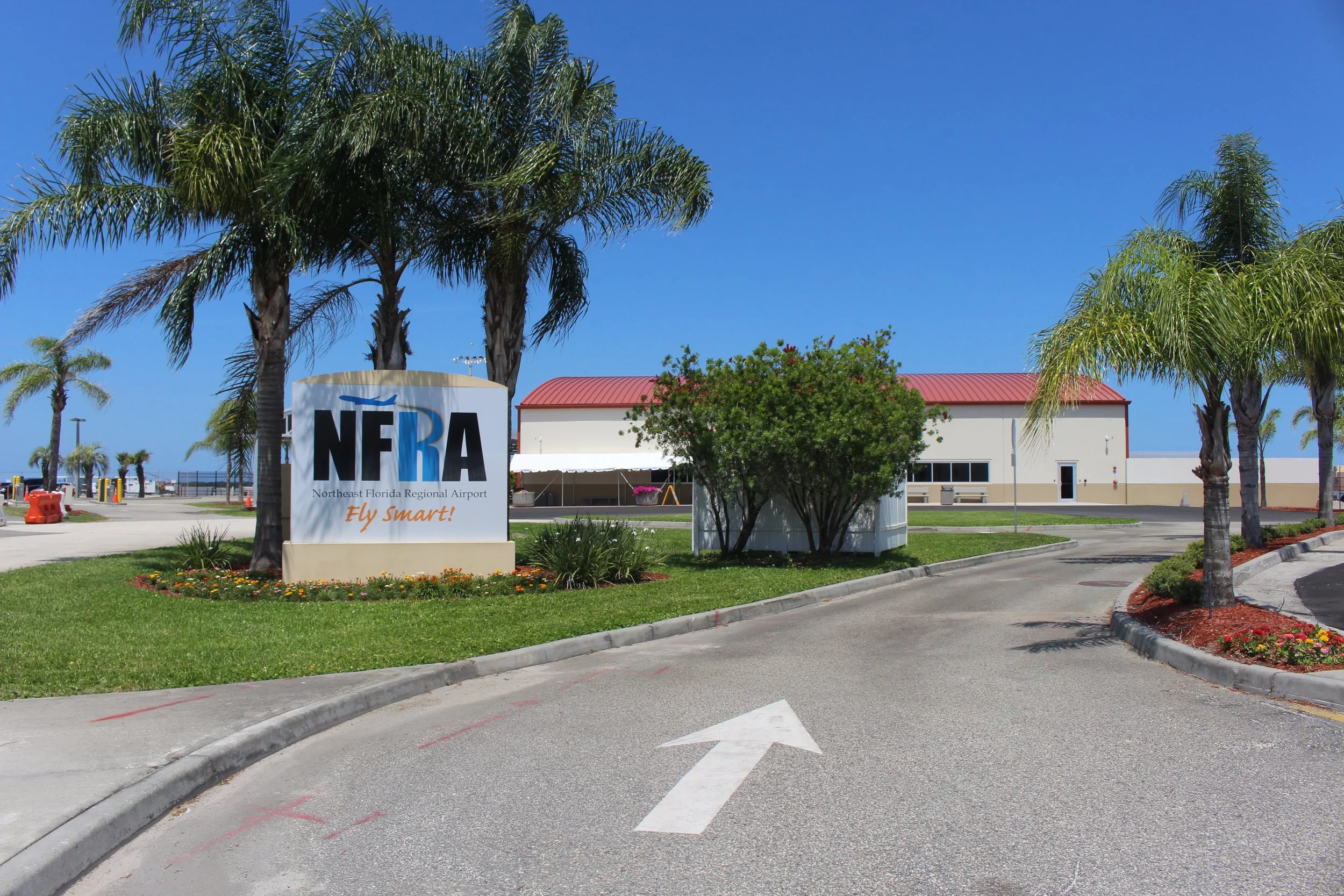 The image shows the entrance to Northeast Florida Regional Airport with palm trees, a sign that reads 'NFRA' and 'Fly Smart!', a building with a red roof, and a clear blue sky.