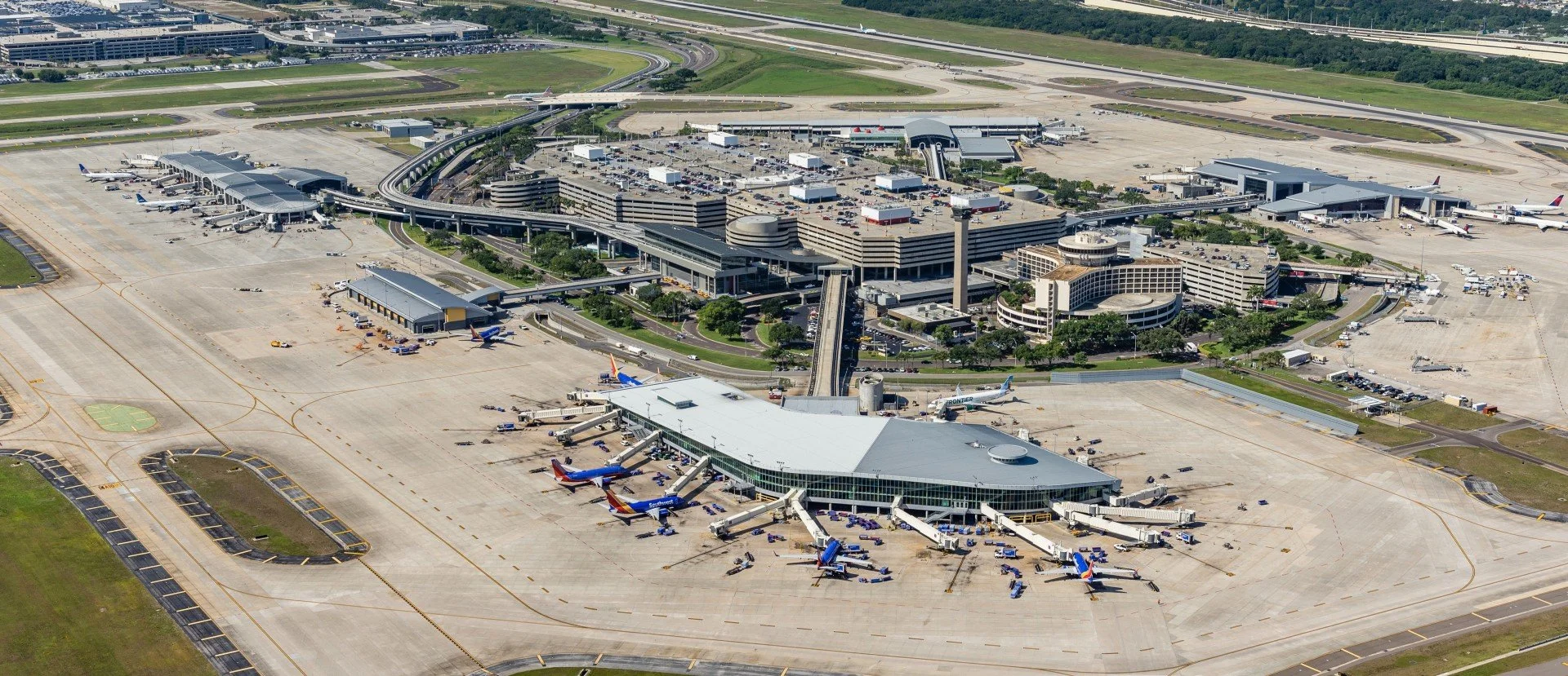 Aerial view of an airport terminal with multiple airplanes parked at gates, jet bridges connected to planes, and a large parking structure nearby. Runways and taxiways are visible in the background.