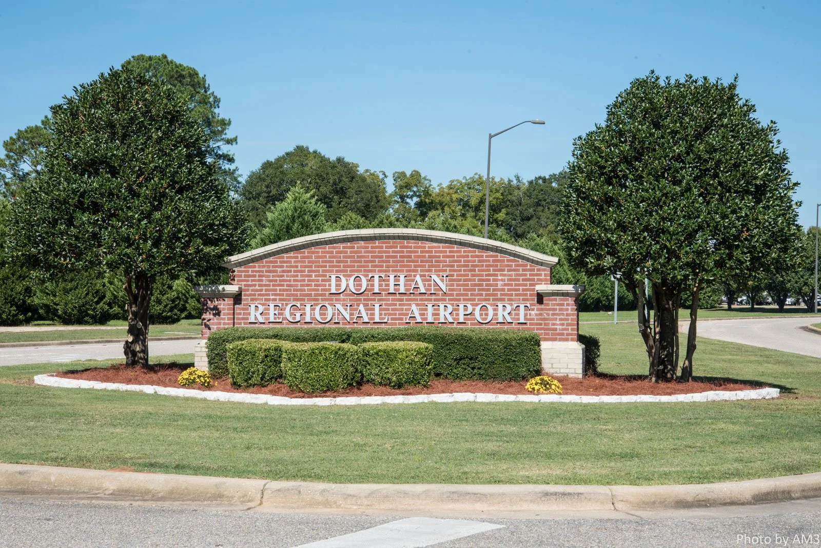 Sign reads 'Dothan Regional Airport' with brick border, surrounded by green bushes and trees on a clear day.
