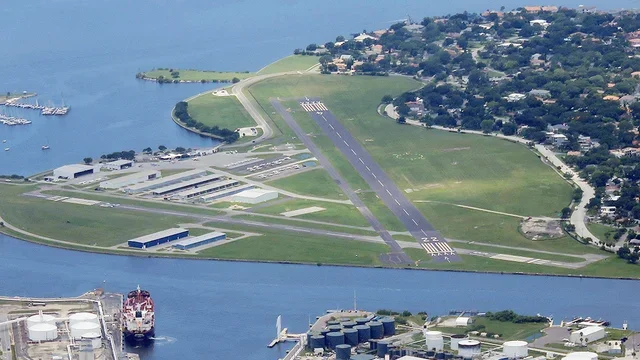 Aerial view of a small airport with a single runway, taxiways, and surrounding water, neighborhoods, and industrial area.