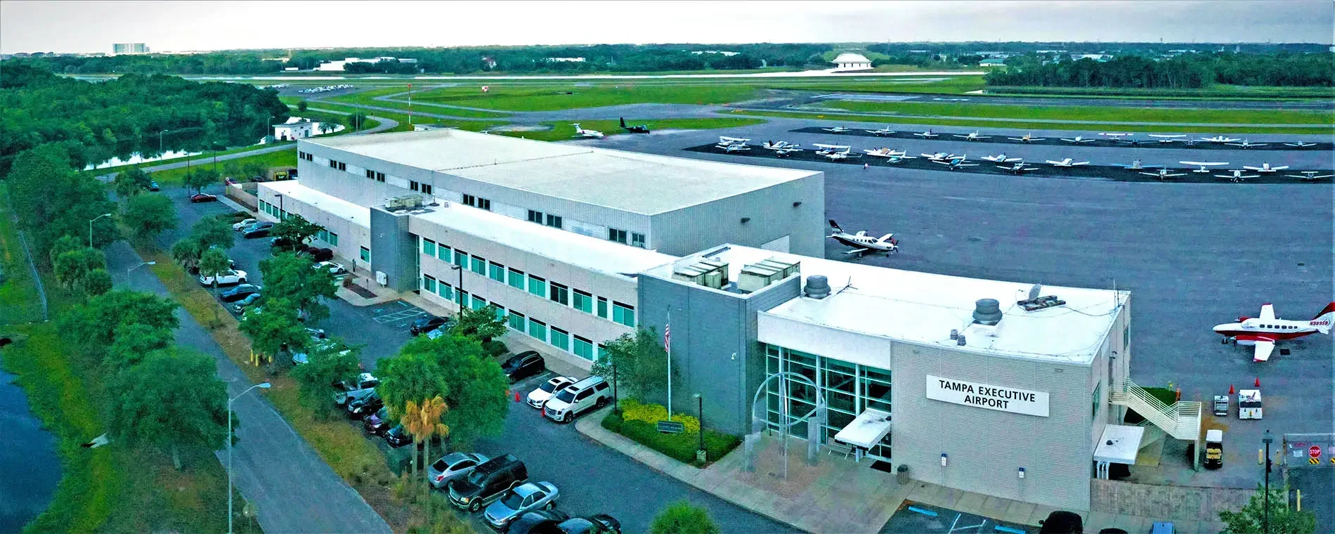 Aerial view of Tampa Executive Airport showing a terminal building with parking lot, several small airplanes parked on the tarmac, and a runway in the background surrounded by green trees and open fields.