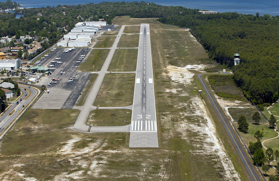 Aerial view of a small regional airport with a single runway, taxiways, and airport buildings, green areas, and a body of water in the background.