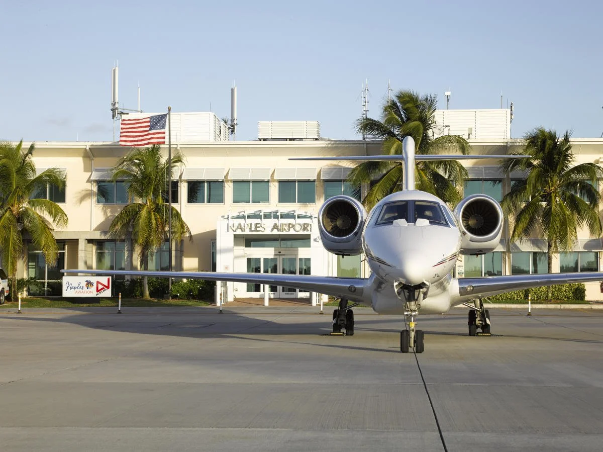 Front view of a private jet parked in front of Naples Airport terminal building, with palm trees and an American flag flying nearby.