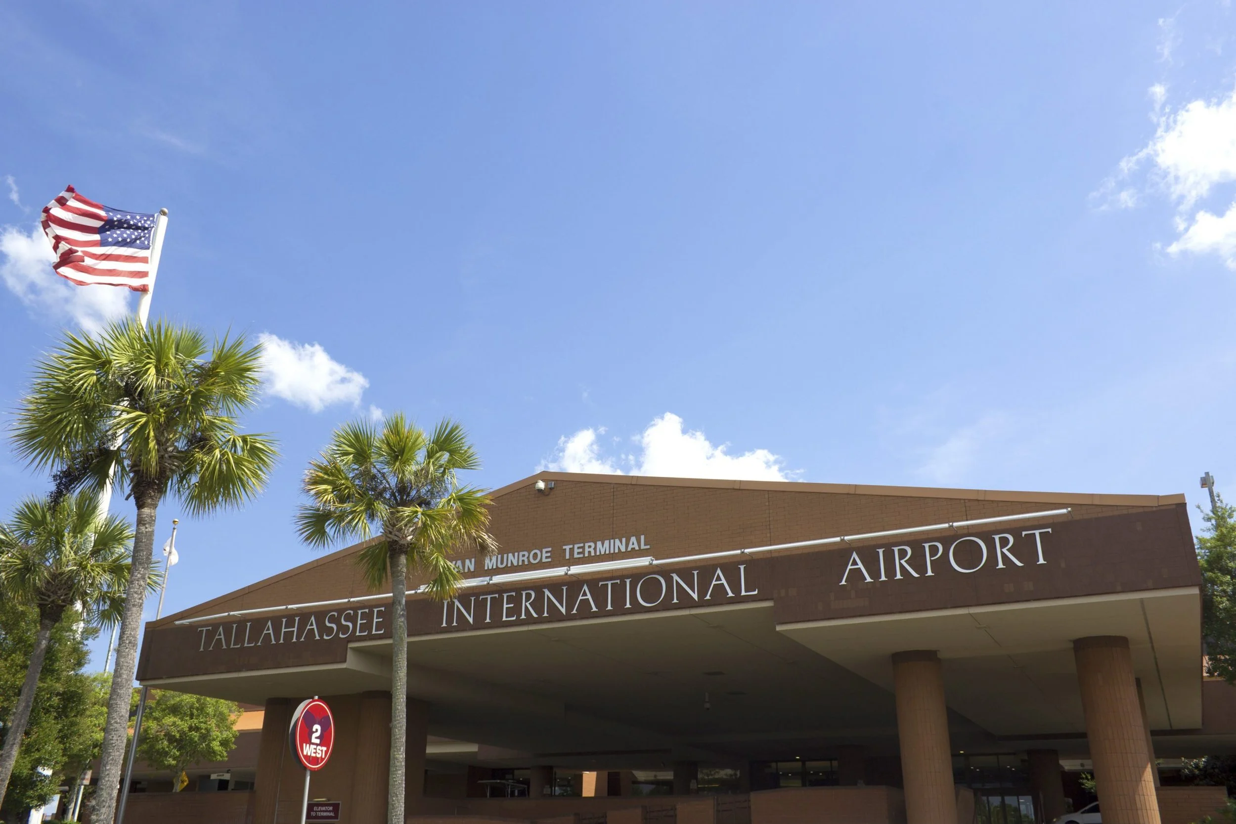 American flag flying above Tallahassee International Airport with palm trees and part of the airport building visible.