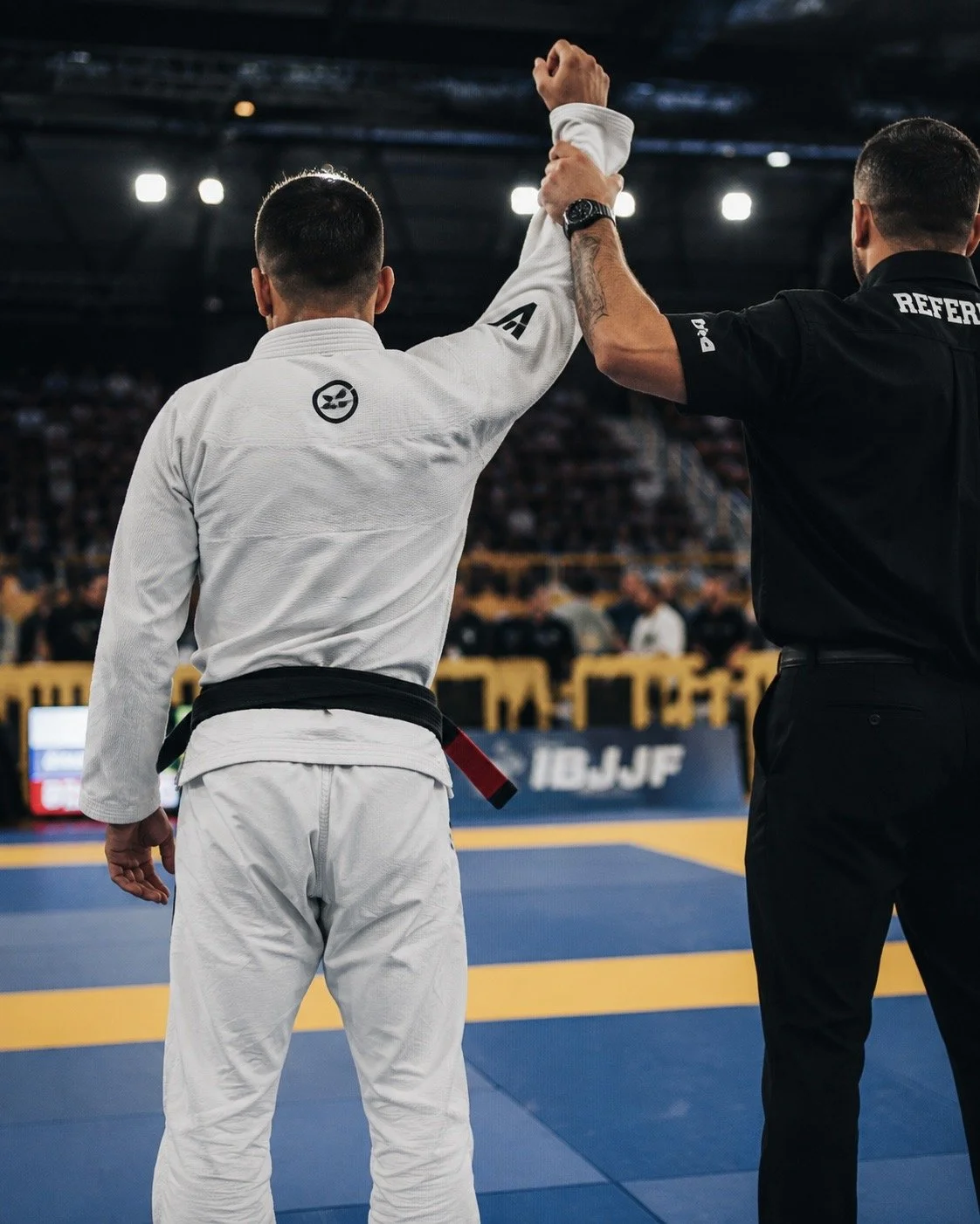 A judo match in progress showing a competitor in a white judogi with a black belt and a referee in a black uniform on a blue and yellow mat, inside a large indoor arena.