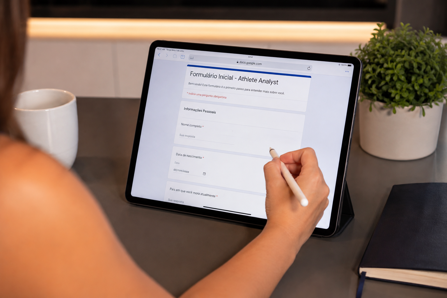 Person filling out an online athlete analyst application form on a tablet with a stylus, sitting at a desk with a notebook, a white cup, and a green plant.