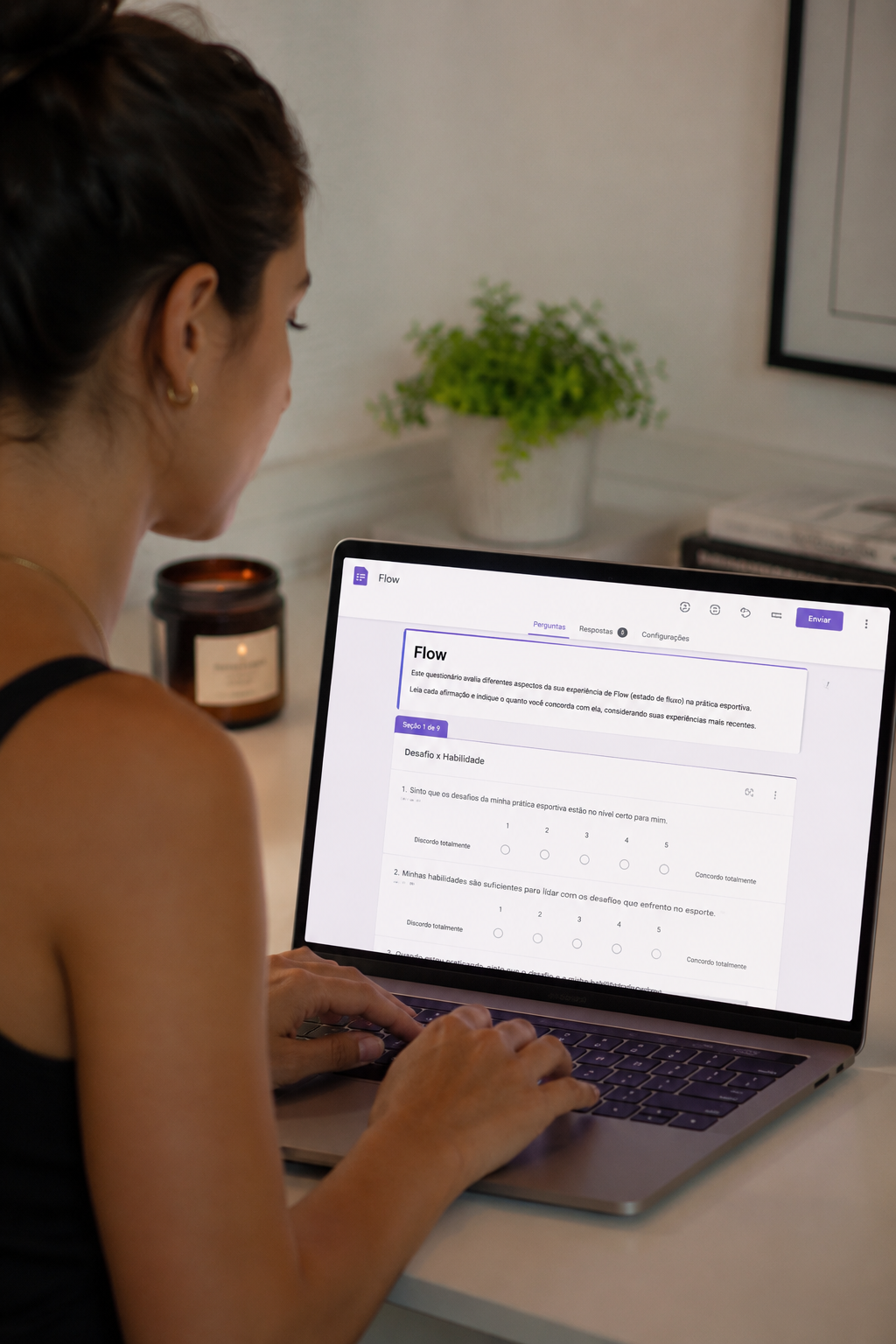 A woman sitting at a white desk using a laptop, viewing a questionnaire titled 'Flow' on the screen in Portuguese, with a background of a small plant, a candle, and some papers.