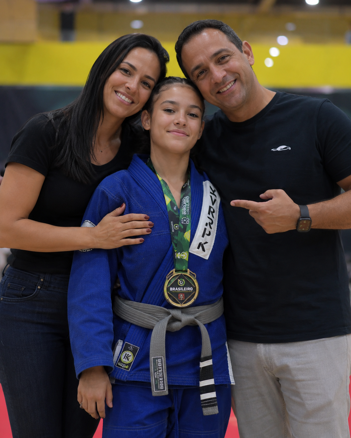 A young girl in a blue judo uniform with a medal around her neck, flanked by a woman and a man, all smiling and posing for the photo.
