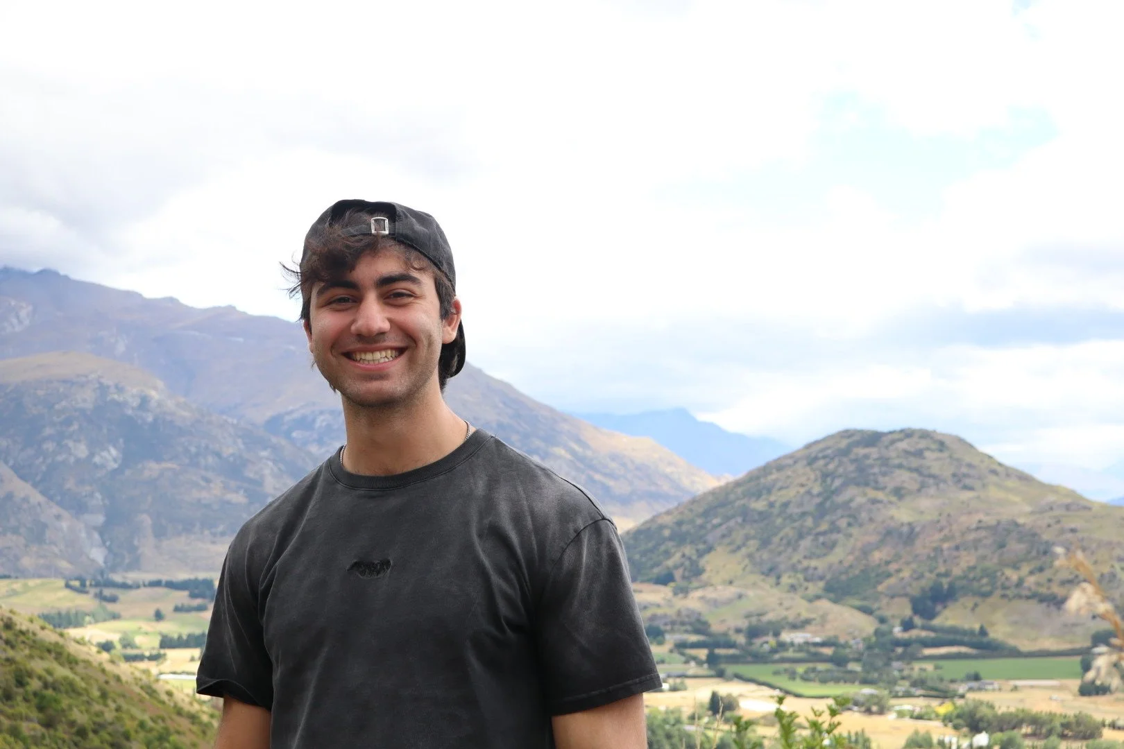 A young man is smiling outdoors with a mountainous landscape in the background. He wears a black t-shirt and a backward baseball cap.