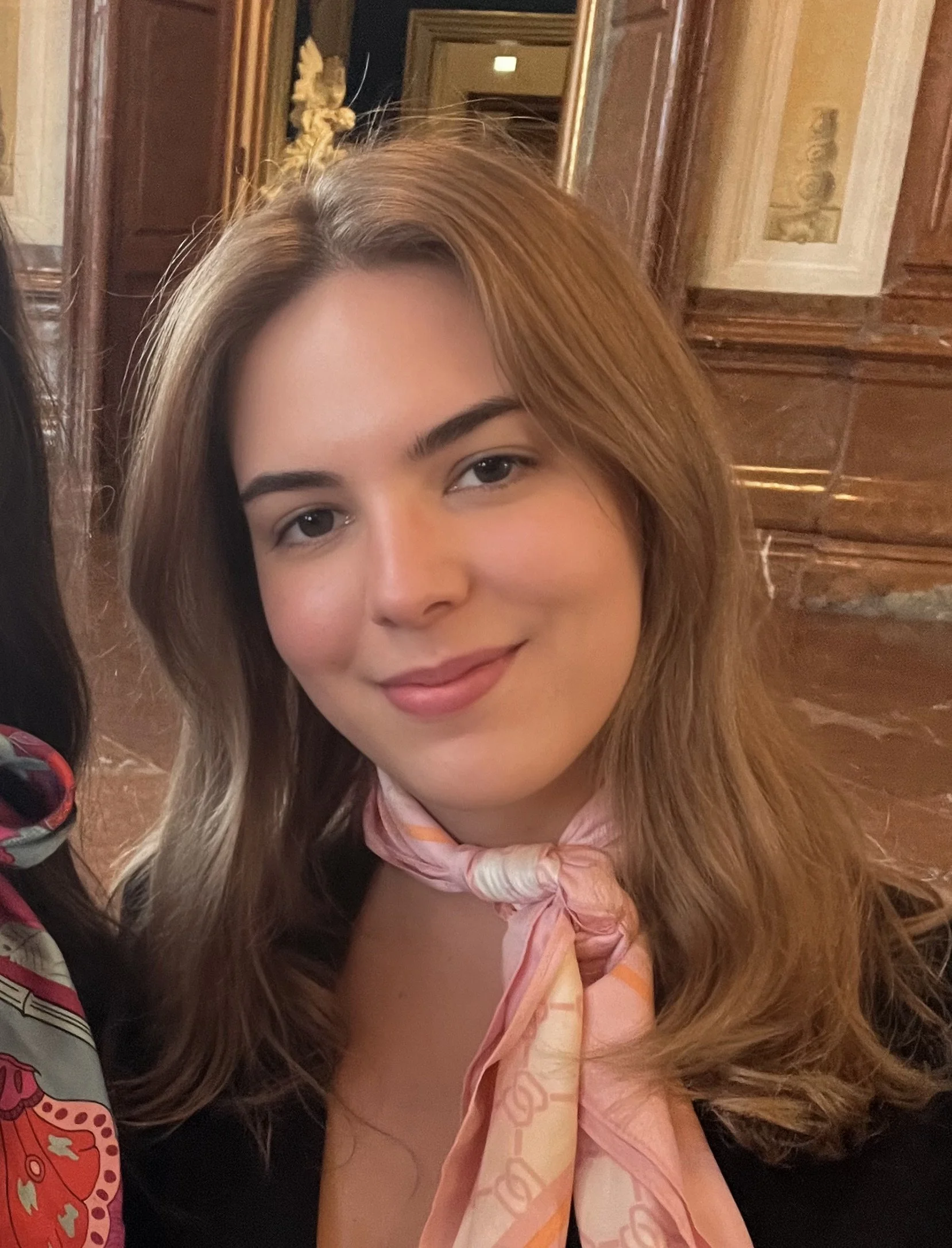 A woman with light skin and long, light brown hair styled in loose waves, smiling softly. She is wearing a pink scarf with orange patterns around her neck. The background features ornate wooden paneling and marble details, indicating an elegant indoor setting.