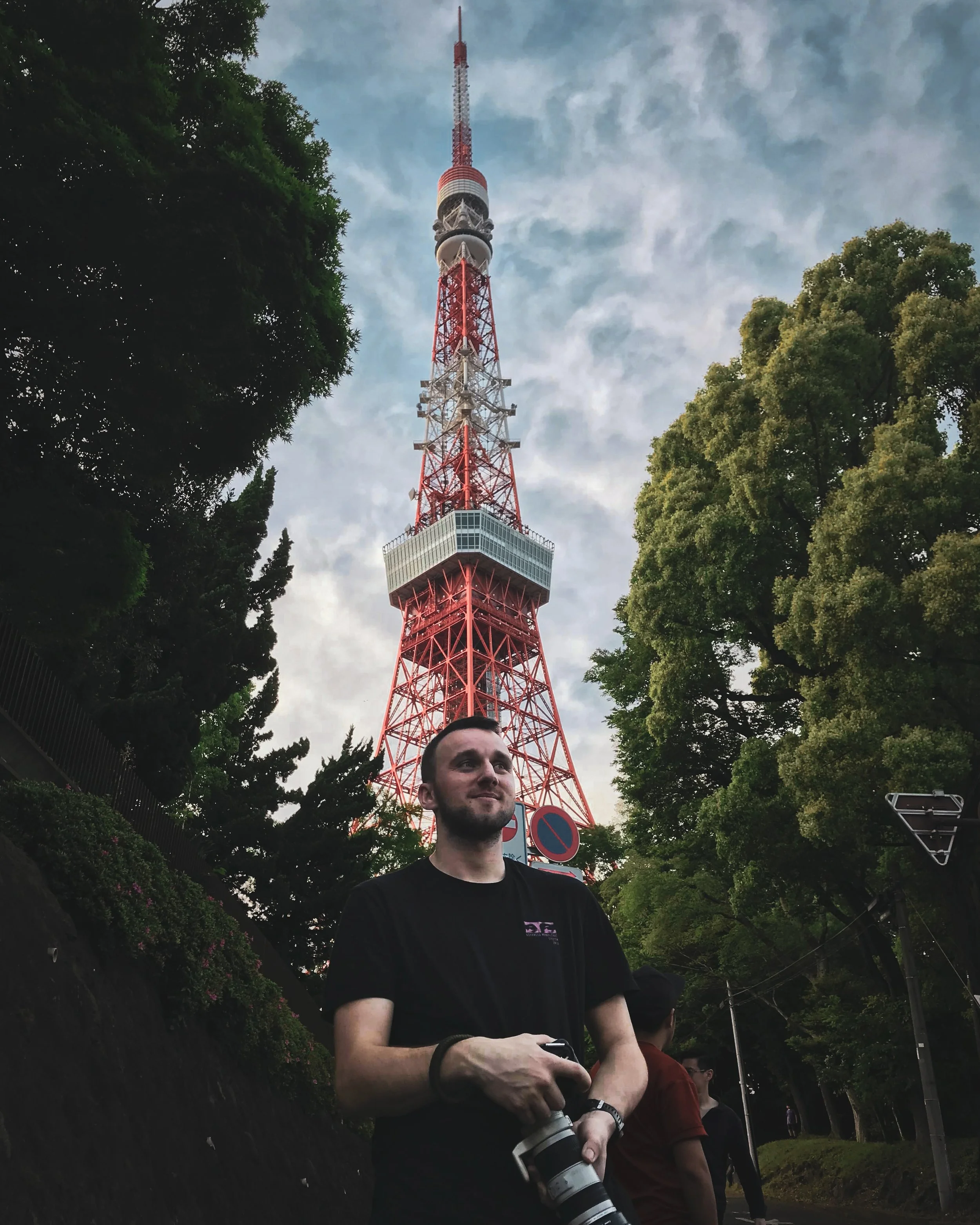 A man with short dark hair and a beard wearing a black t-shirt holding a camera, standing outdoors near trees with a Tokyo Tower in the background.