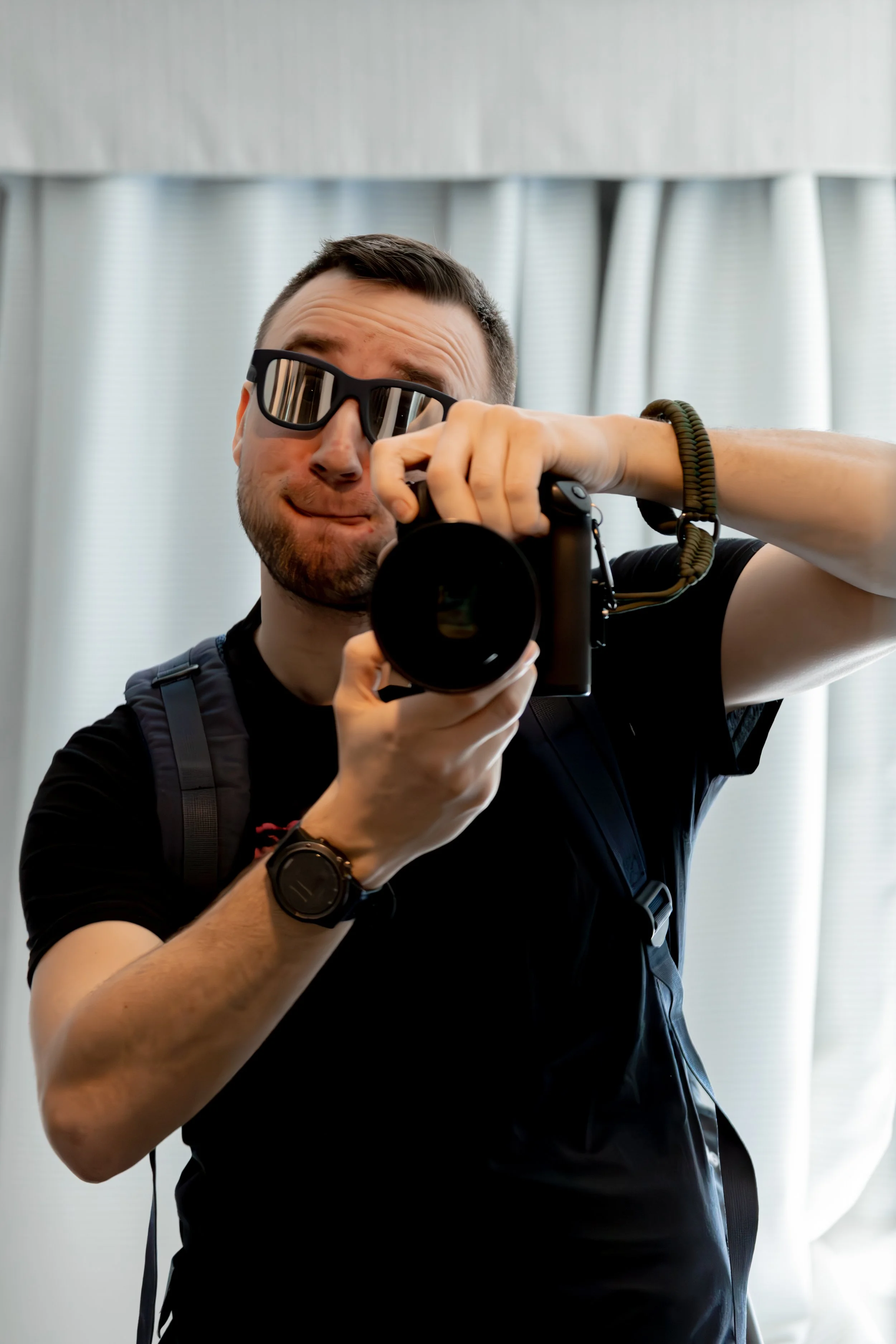 A young man with short dark hair and a beard wearing sunglasses, a black t-shirt, and a watch on his left wrist, taking a selfie with a camera in front of a light-colored curtain.