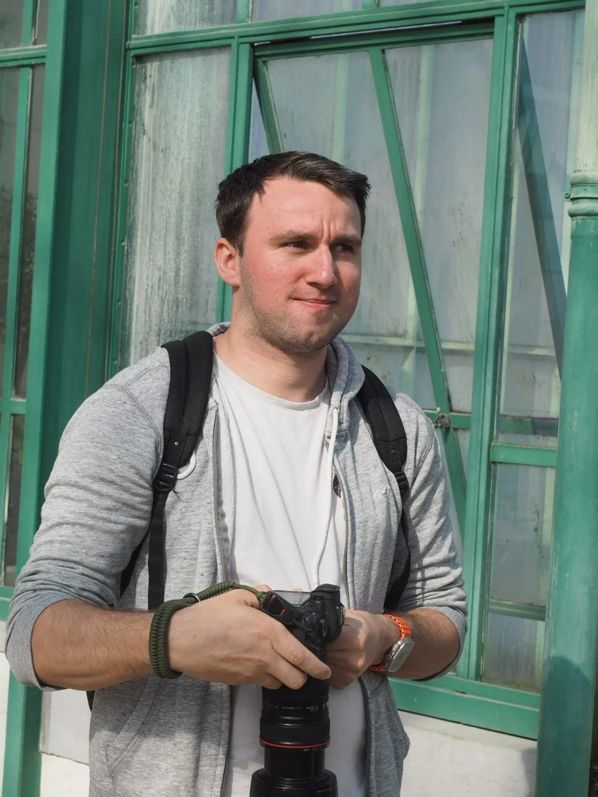A man with short dark hair, wearing a white t-shirt, gray hoodie, and backpack, holding a camera and standing in front of a green structure, possibly a greenhouse.