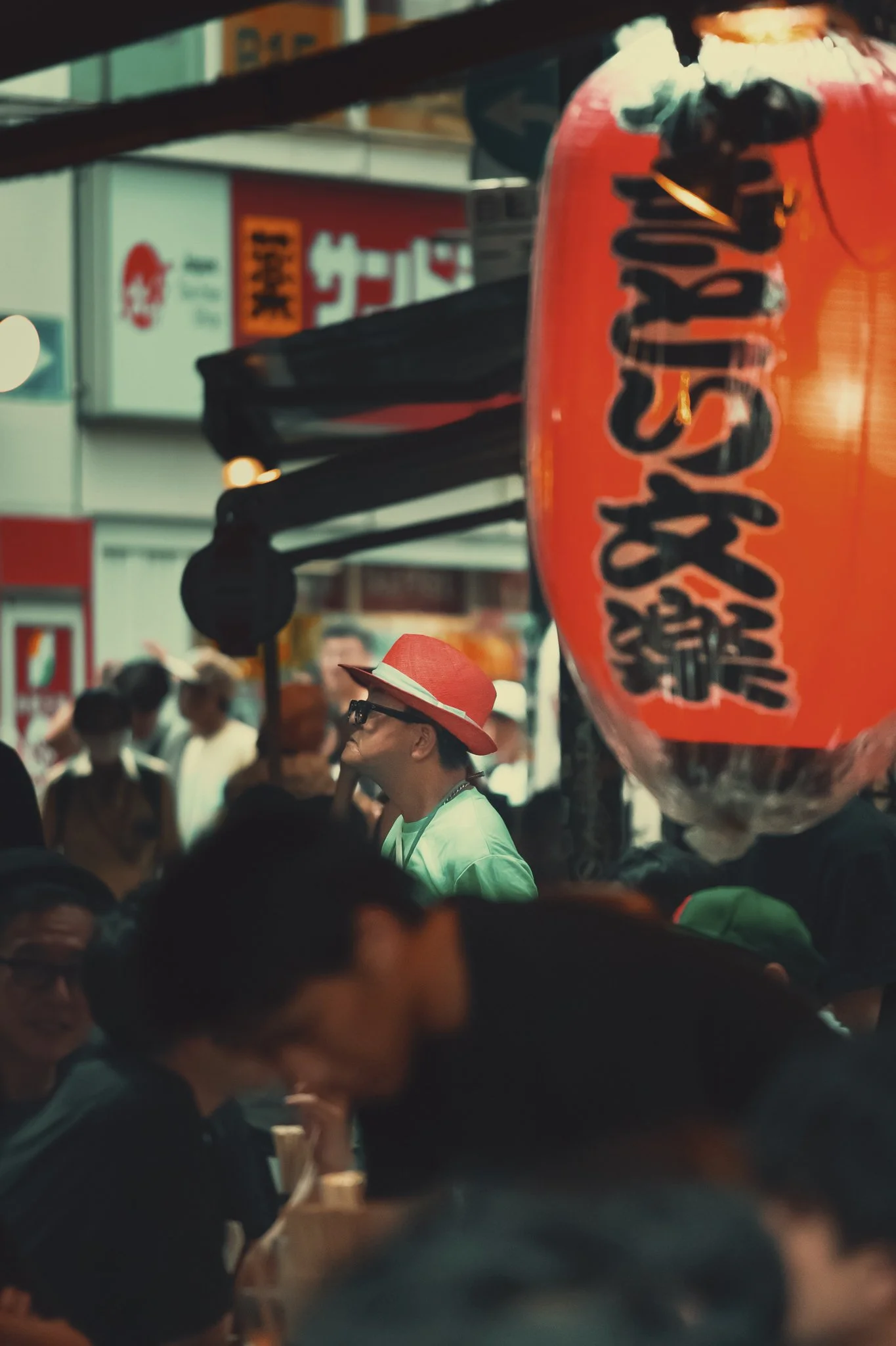 A crowded street scene in Japan with people walking, one wearing a red hat, and a large red lantern with Japanese characters hanging above.