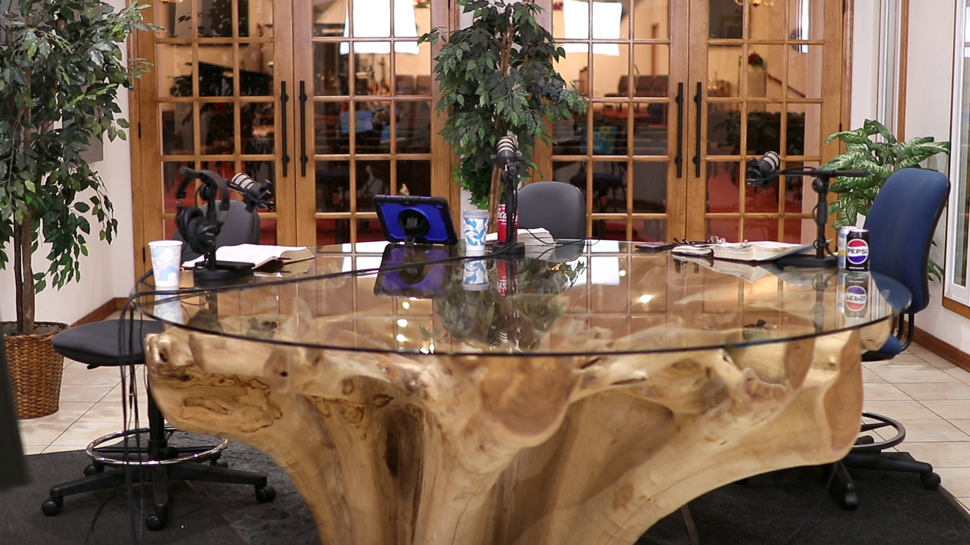 A large, rustic, wooden conference table with four microphones, a tablet, paper plates, cups, glasses, a soda can, and a fire extinguisher. Chairs are positioned around the table, and there are potted plants on either side, with a glass-paneled door and an adjacent window in the background, leading to a room with a sofa and decorations.