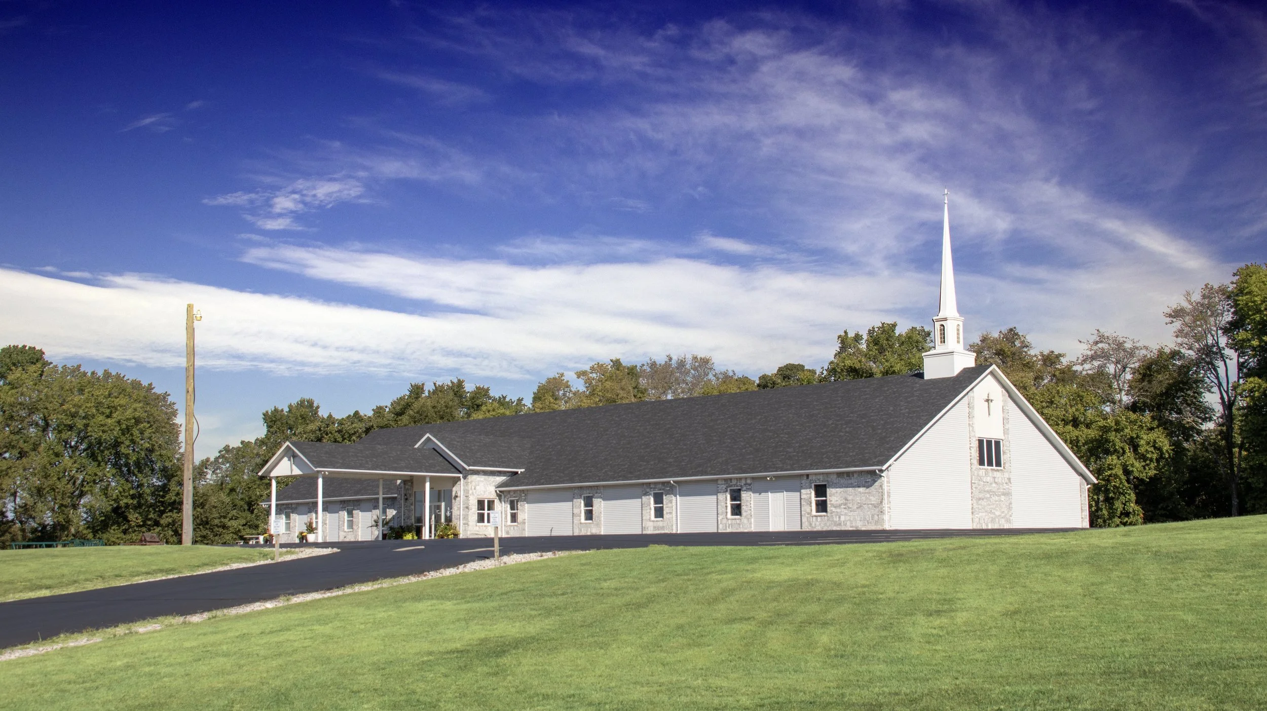 A white church with a tall steeple surrounded by green trees and grass, under a partly cloudy blue sky with light clouds.