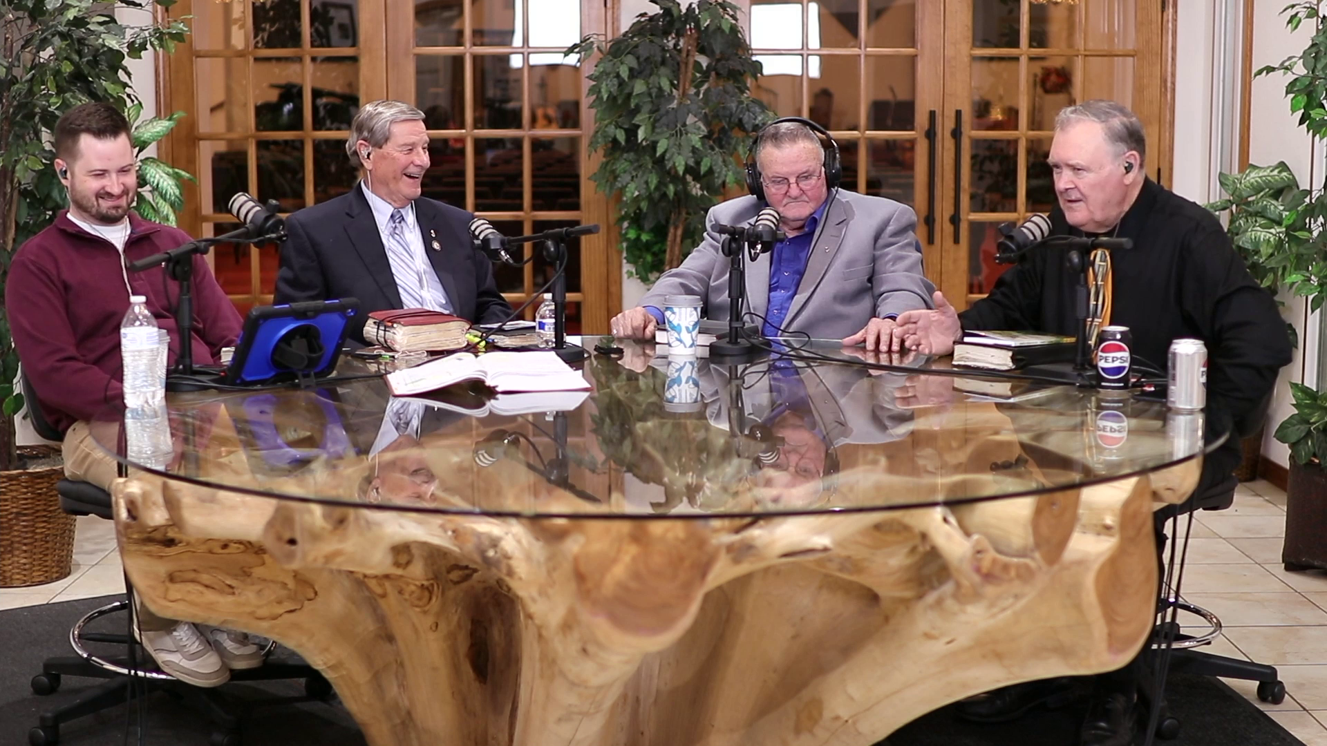 Four men sitting at a large, uniquely shaped wooden table in a room with wooden doors and green plants. They are having a conversation with microphones in front of them, some with notebooks and drinks.