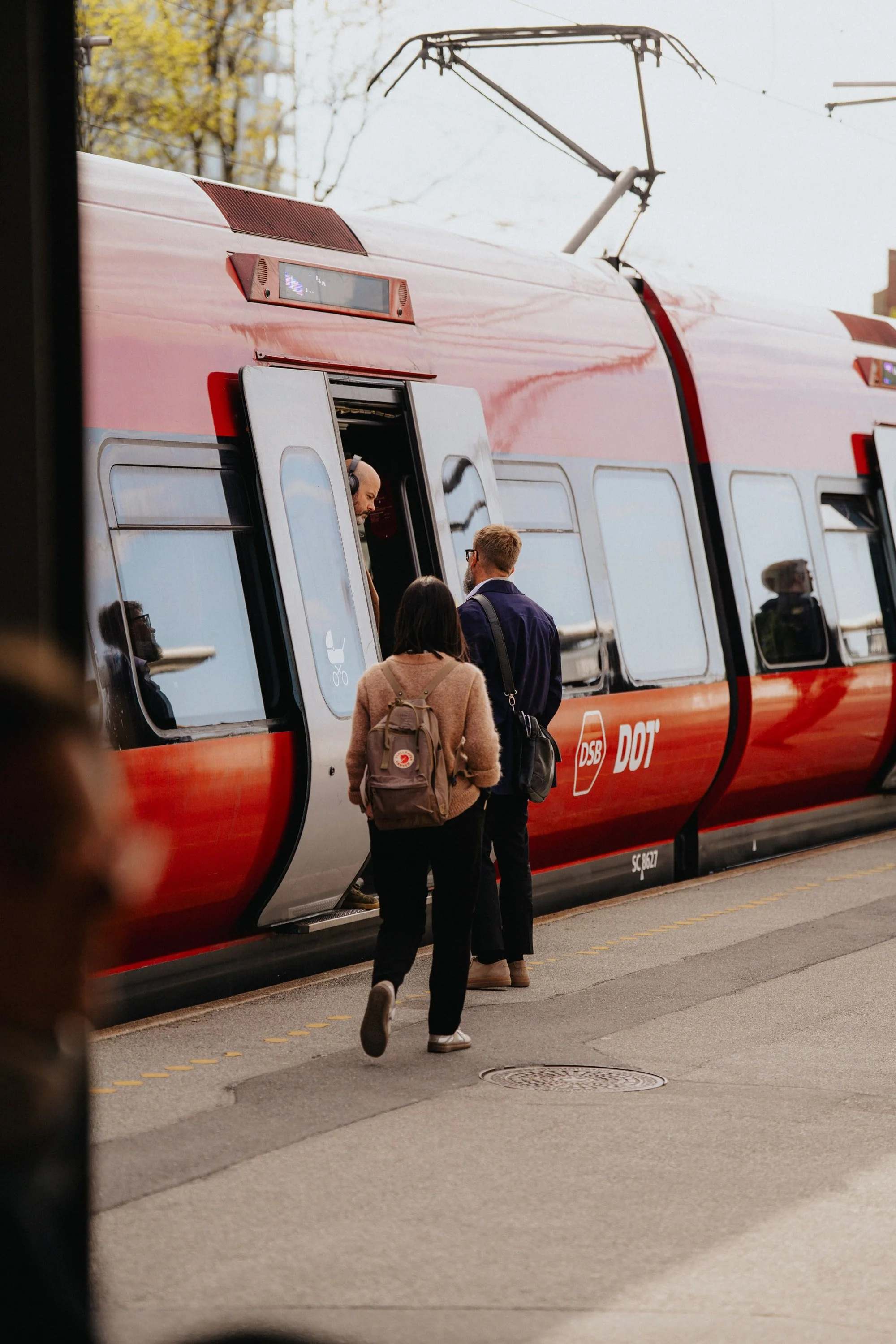 En rød sporvogn med passagerer og chauffør, der står ved døren, mens nogle personer går forbi på fortovet.