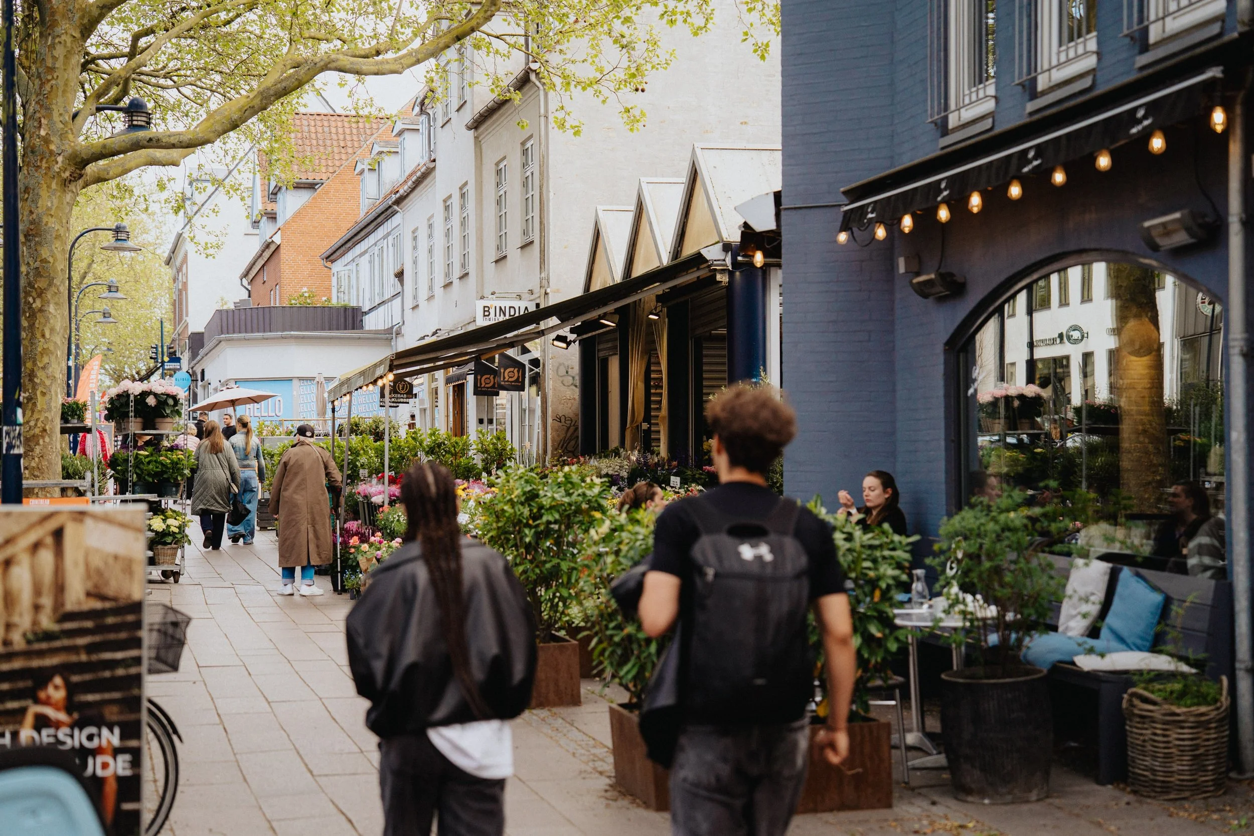Gade med folk, blomsterboder og caféer i en bymidte med træer og bygninger.