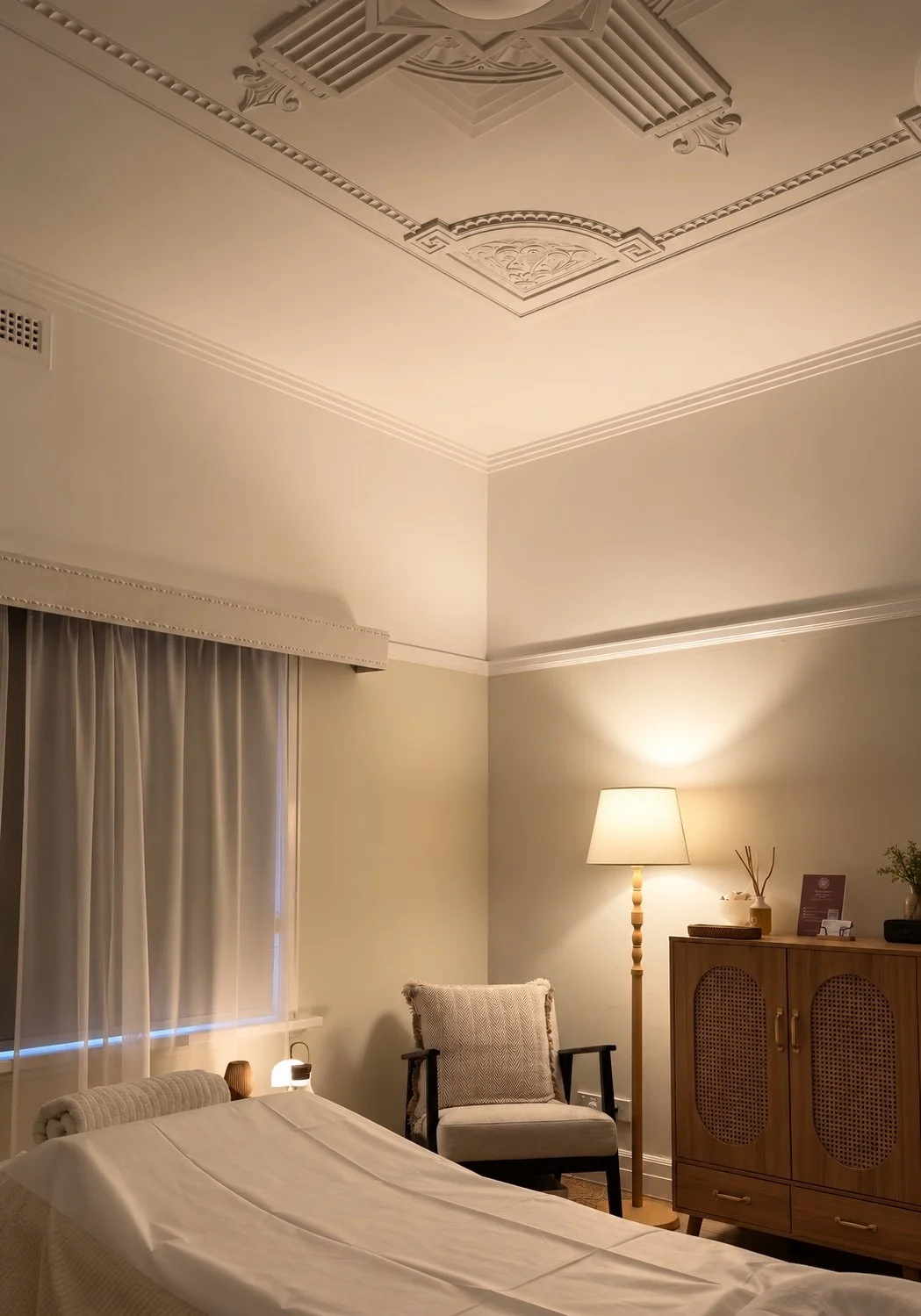 A cozy bedroom with soft lighting, a bed covered with a white sheet, a window with closed curtains, a sitting armchair, a wooden cabinet with decor items, a floor lamp, and decorative ceiling moldings.
